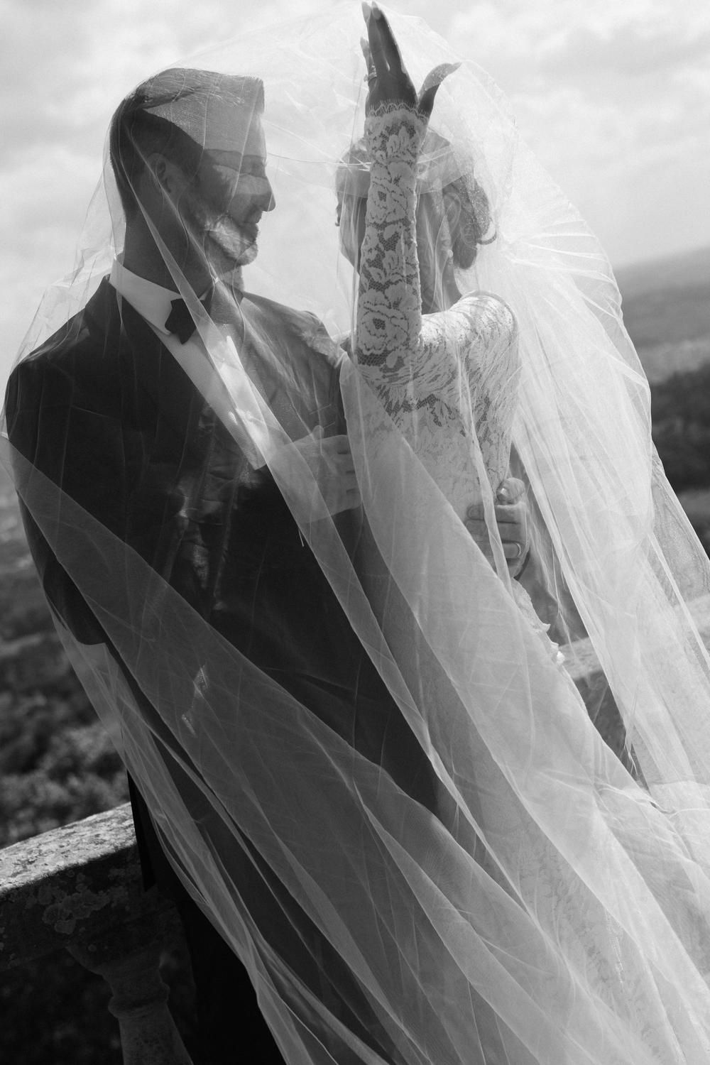 Black and white artistic photo of bride and groom draped together under a flowing veil.