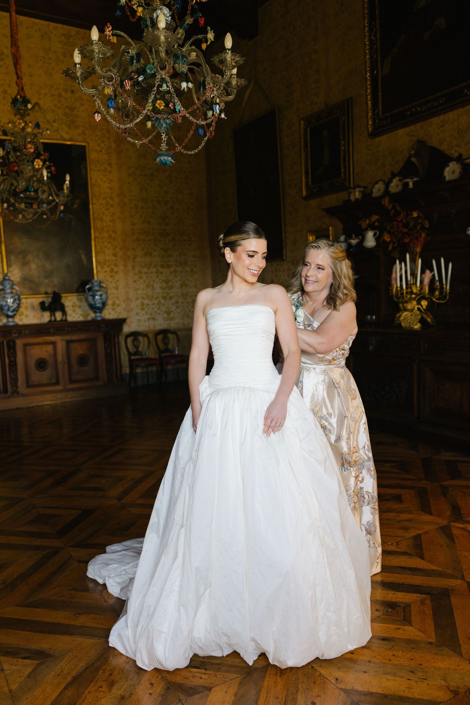 Bride in strapless ball gown being helped by a woman in a gold dress inside an ornate chandelier-lit room.