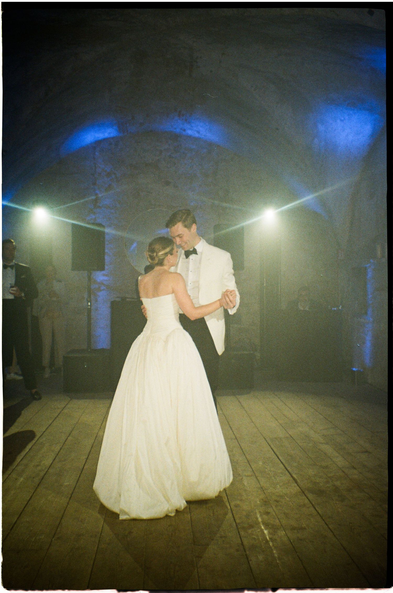 Bride and groom share first dance in stone-vaulted venue with dramatic blue and white stage lighting.
