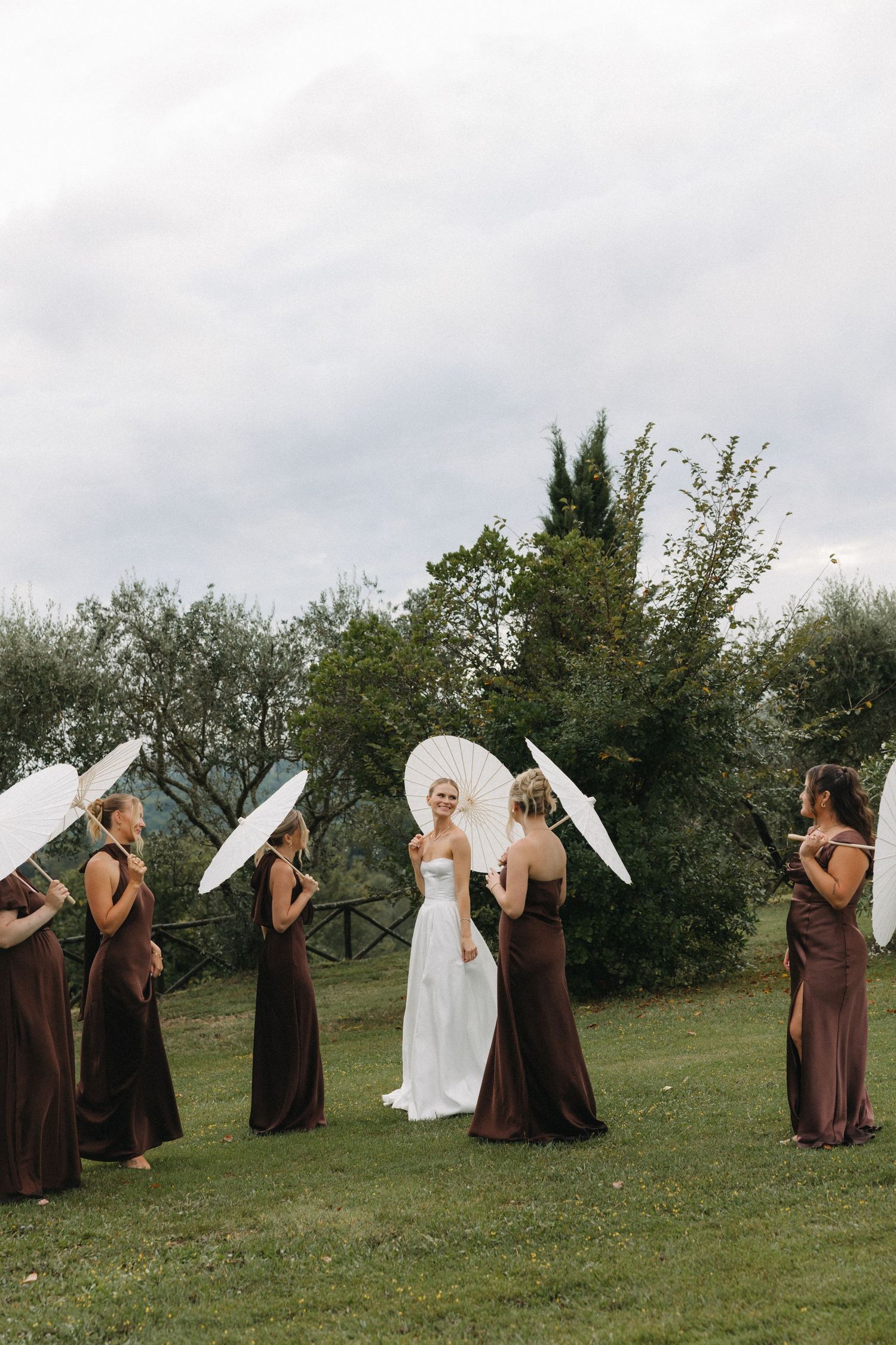 Bride in white gown stands with bridesmaids in brown dresses holding white parasols on green lawn with trees.