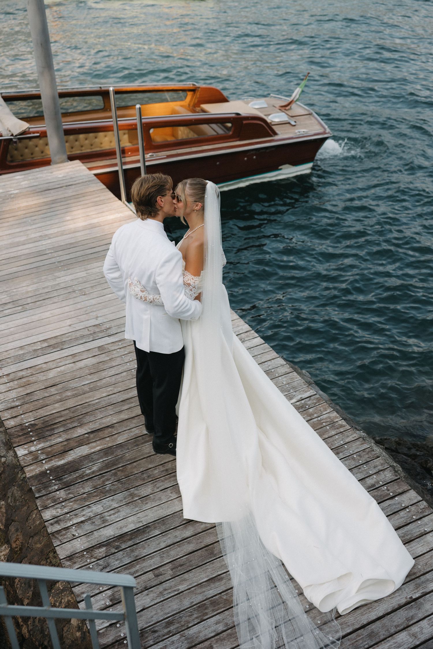 Bride and groom kissing on a wooden dock beside a classic wooden boat on blue water.