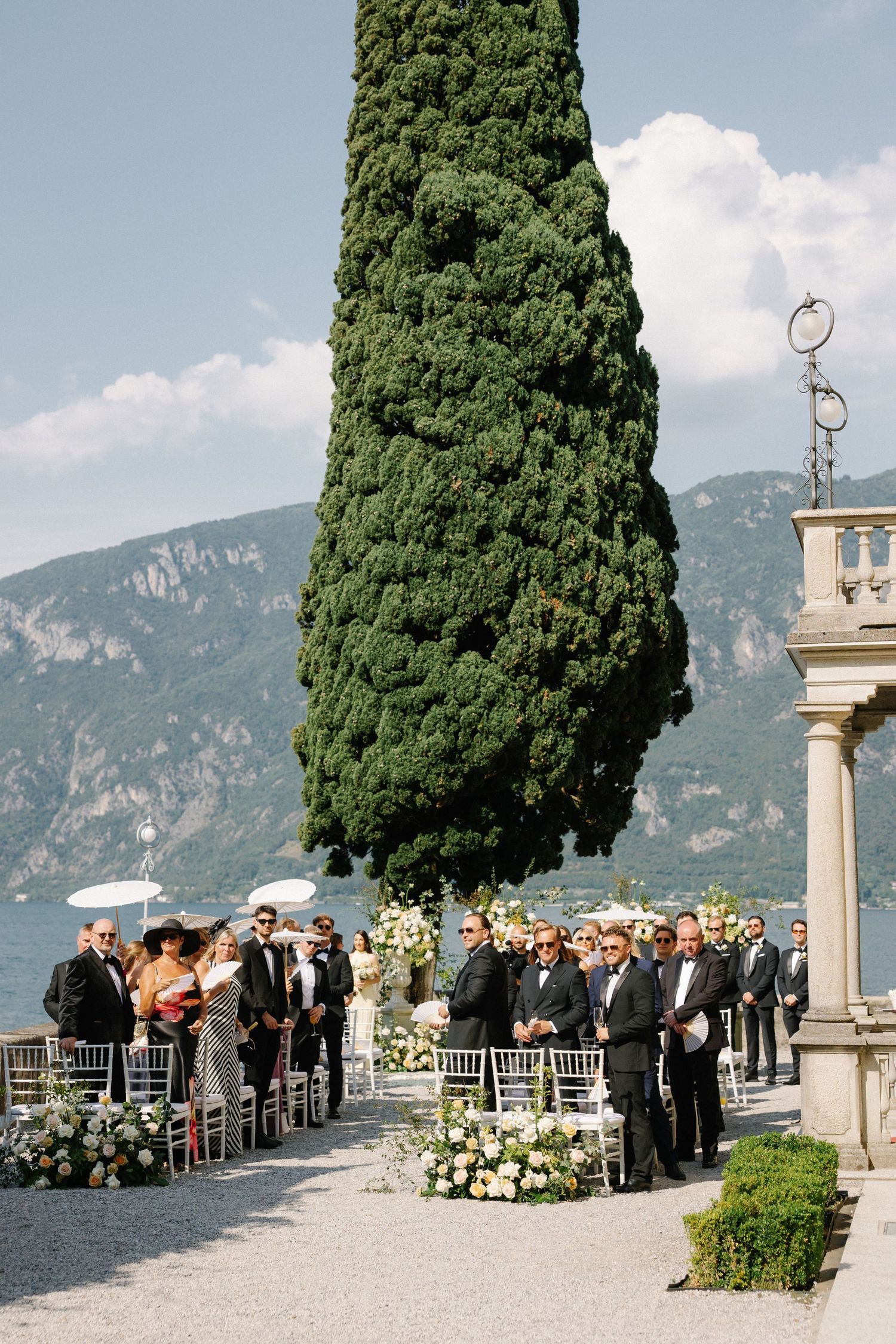 Outdoor wedding ceremony on a lakeside terrace with floral arrangements, cypress tree, and mountain views at Lake Como.