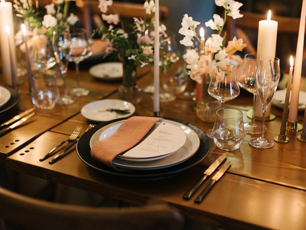 Elegant wedding reception table setting with black plates, gold cutlery, candles, and floral centerpieces.