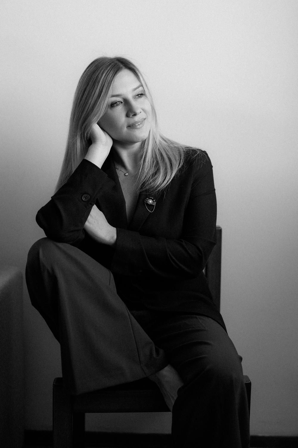 Black and white portrait of blonde woman in black outfit seated on chair with thoughtful expression.