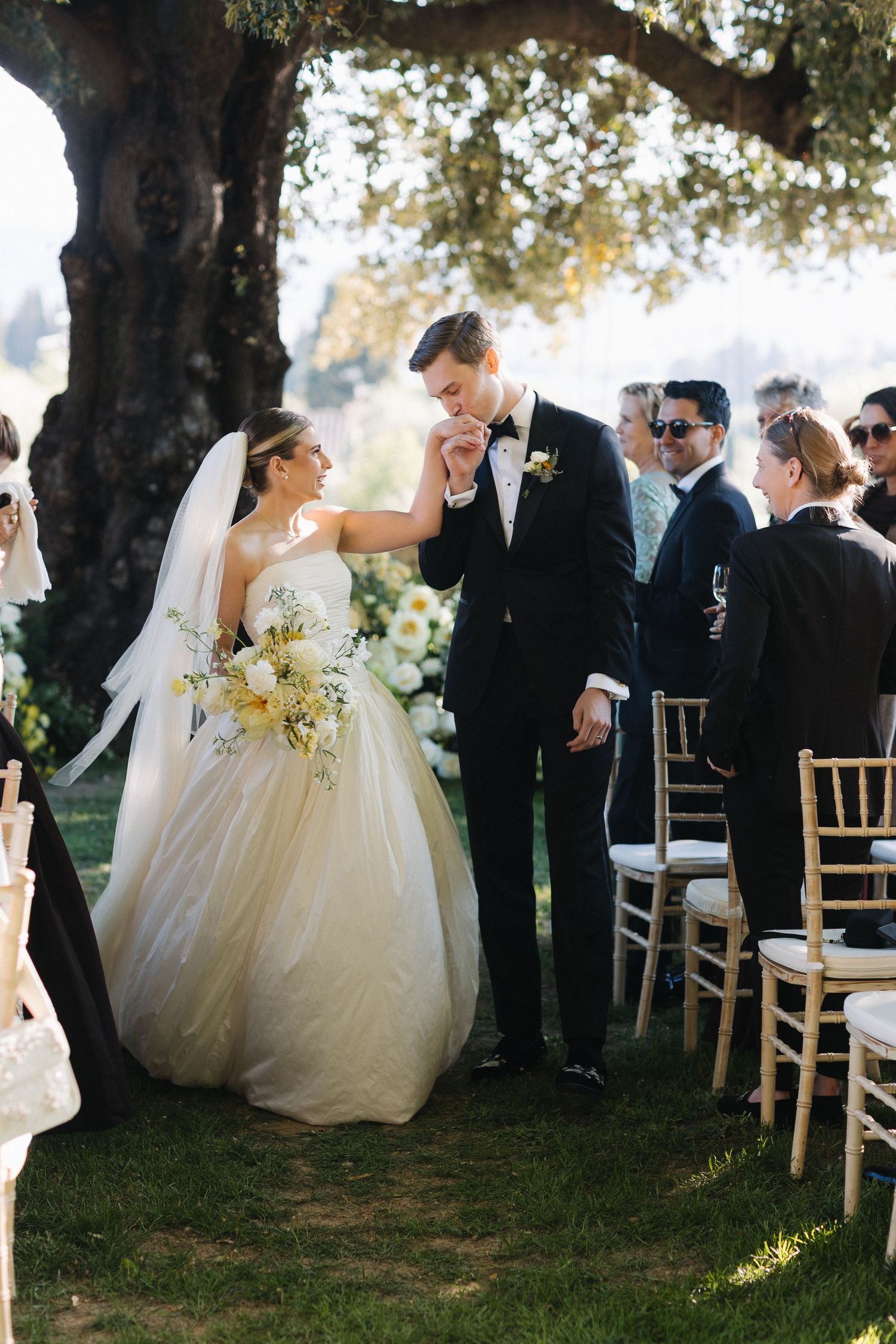 Groom kissing bride's hand during outdoor wedding ceremony under an olive tree with guests seated nearby.