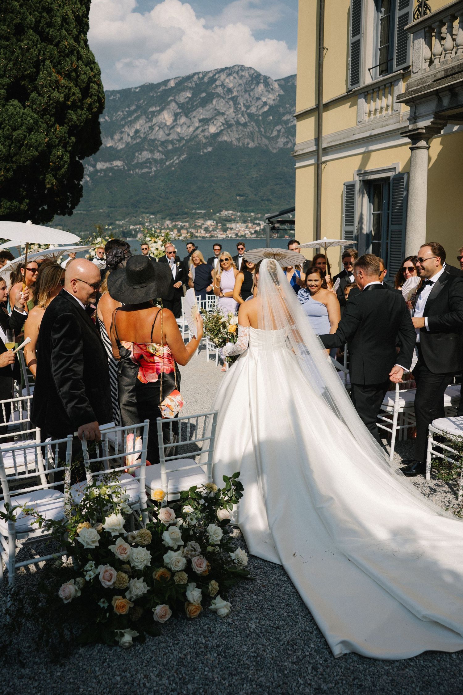 Bride walks down aisle in white gown with long veil at outdoor wedding ceremony with mountain backdrop.