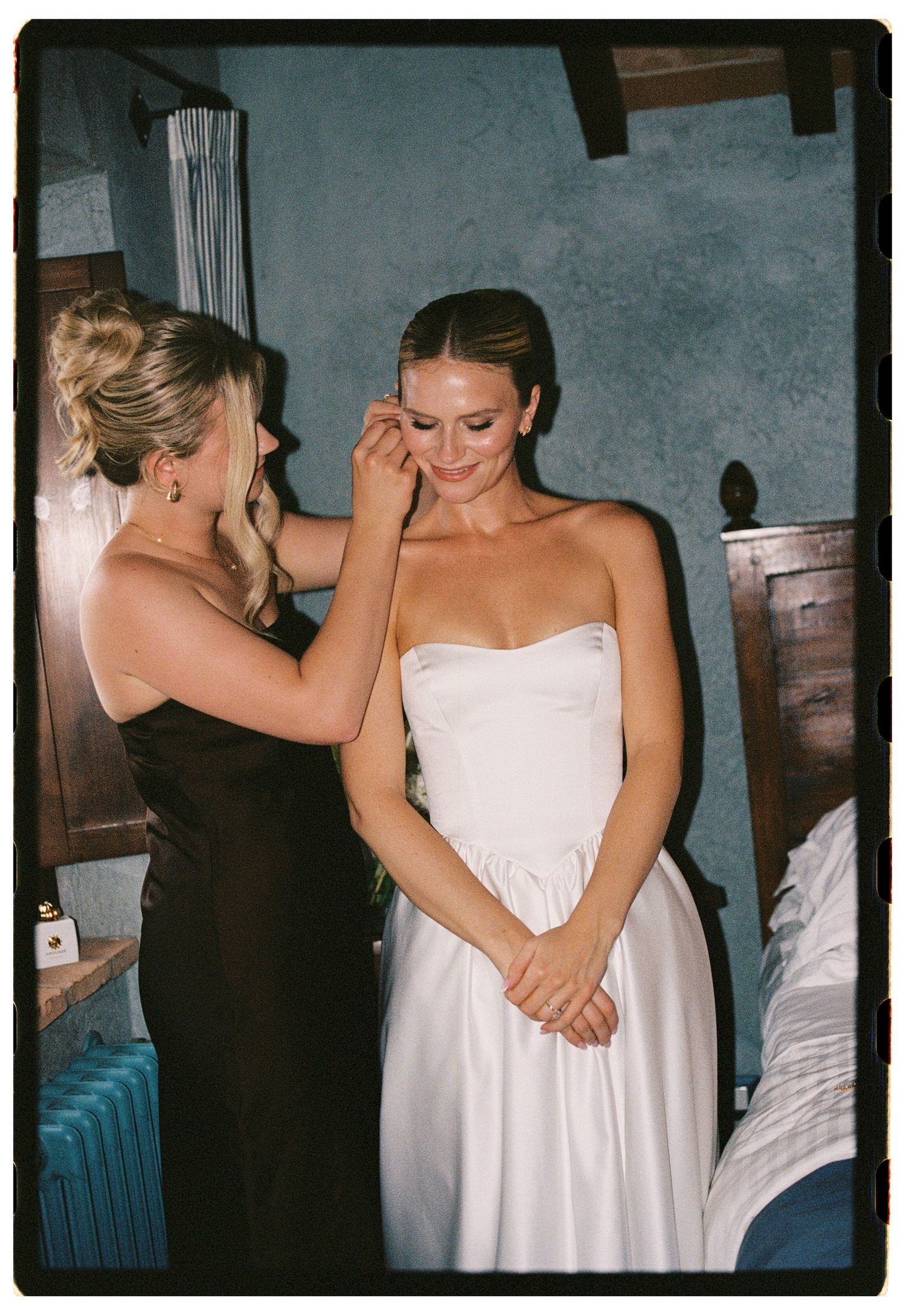 Bride in strapless white gown gets makeup touch-up from bridesmaid in black dress in rustic blue room.
