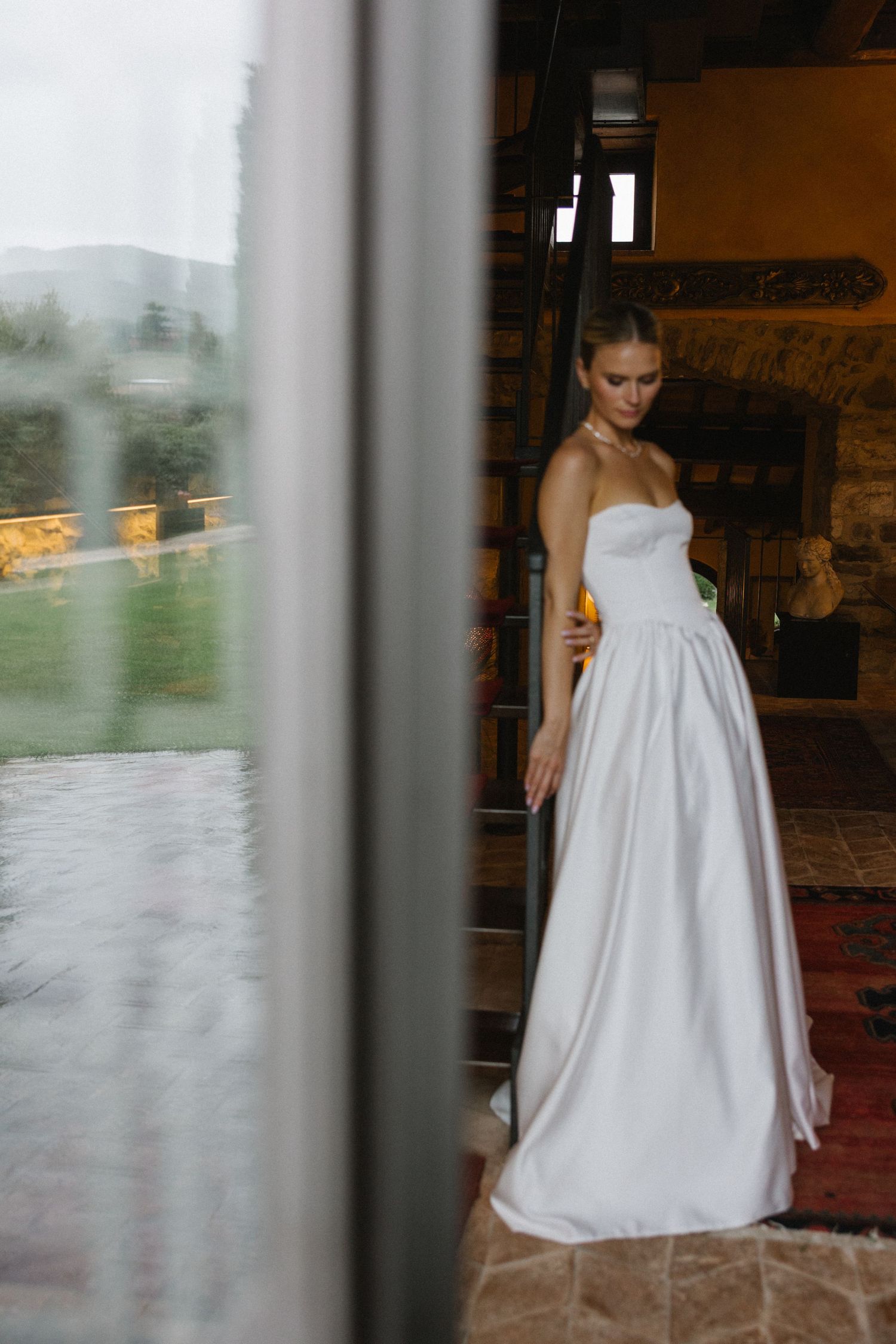 Elegant bride in a strapless white satin ball gown standing in a rustic venue doorway with scenic outdoor views.