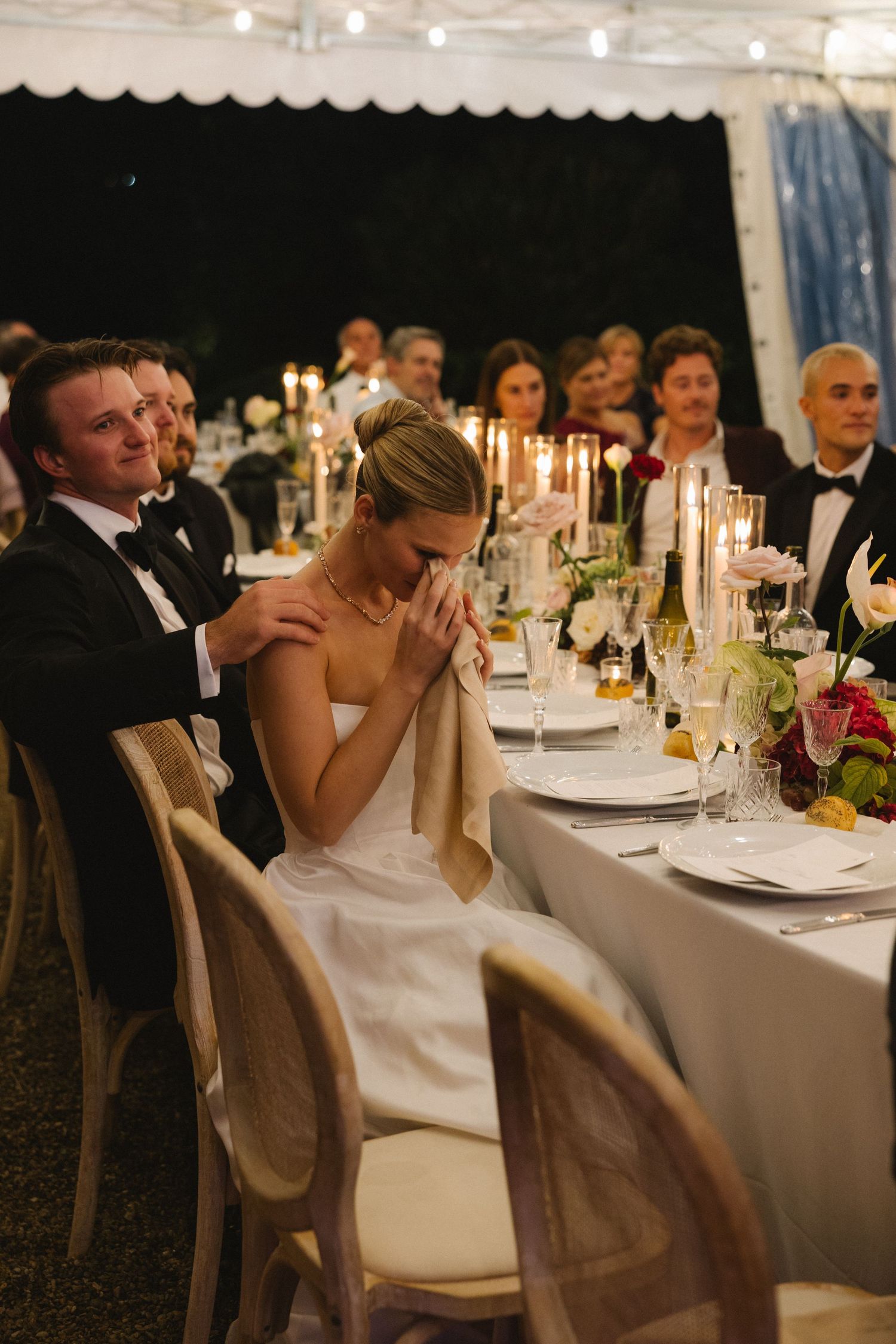 Bride wiping tears at wedding reception dinner table while groom comforts her, surrounded by candlelit decor.