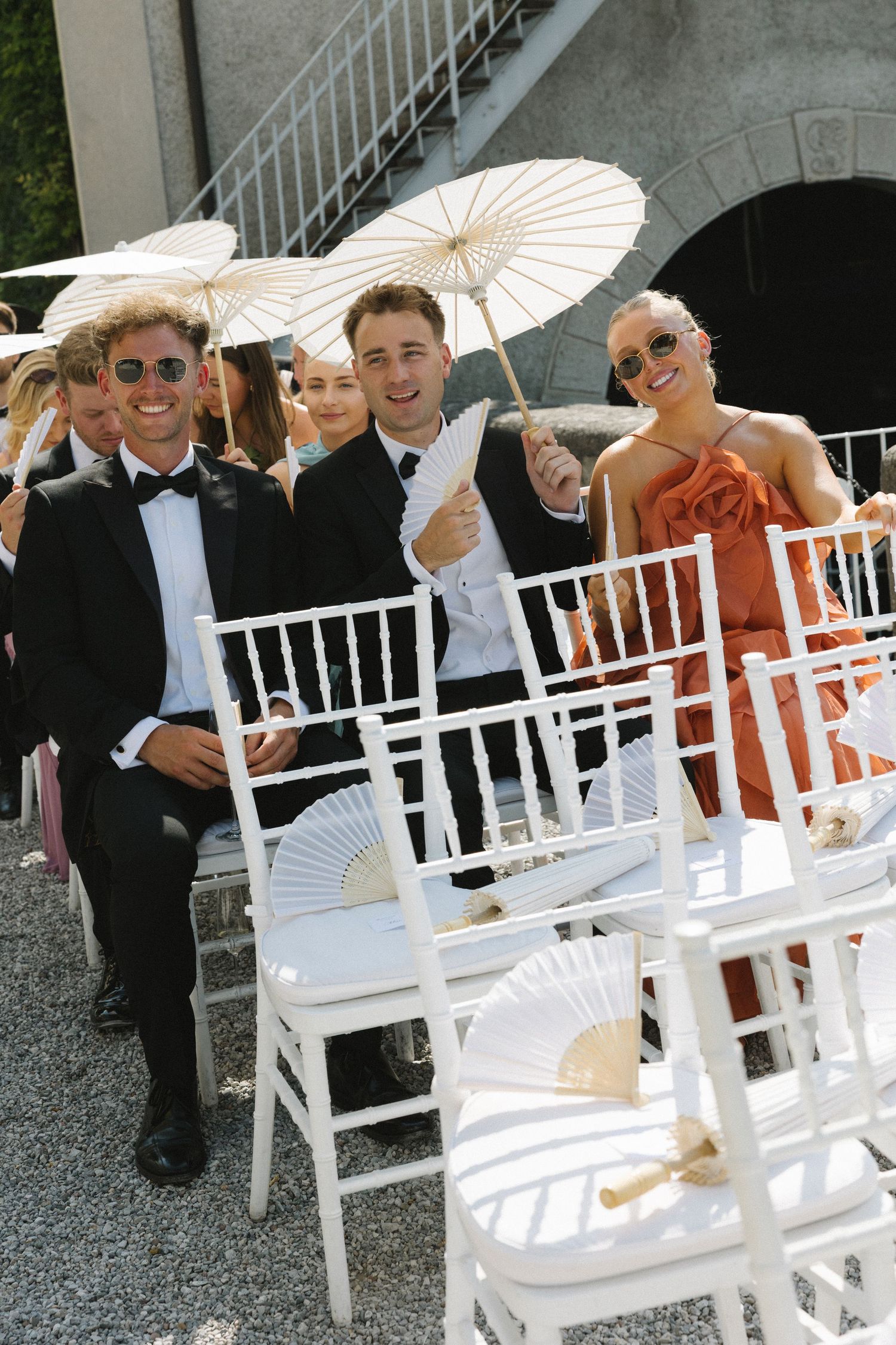 Elegantly dressed wedding guests sit on white chairs, holding parasols and fans outdoors at a sunny ceremony.