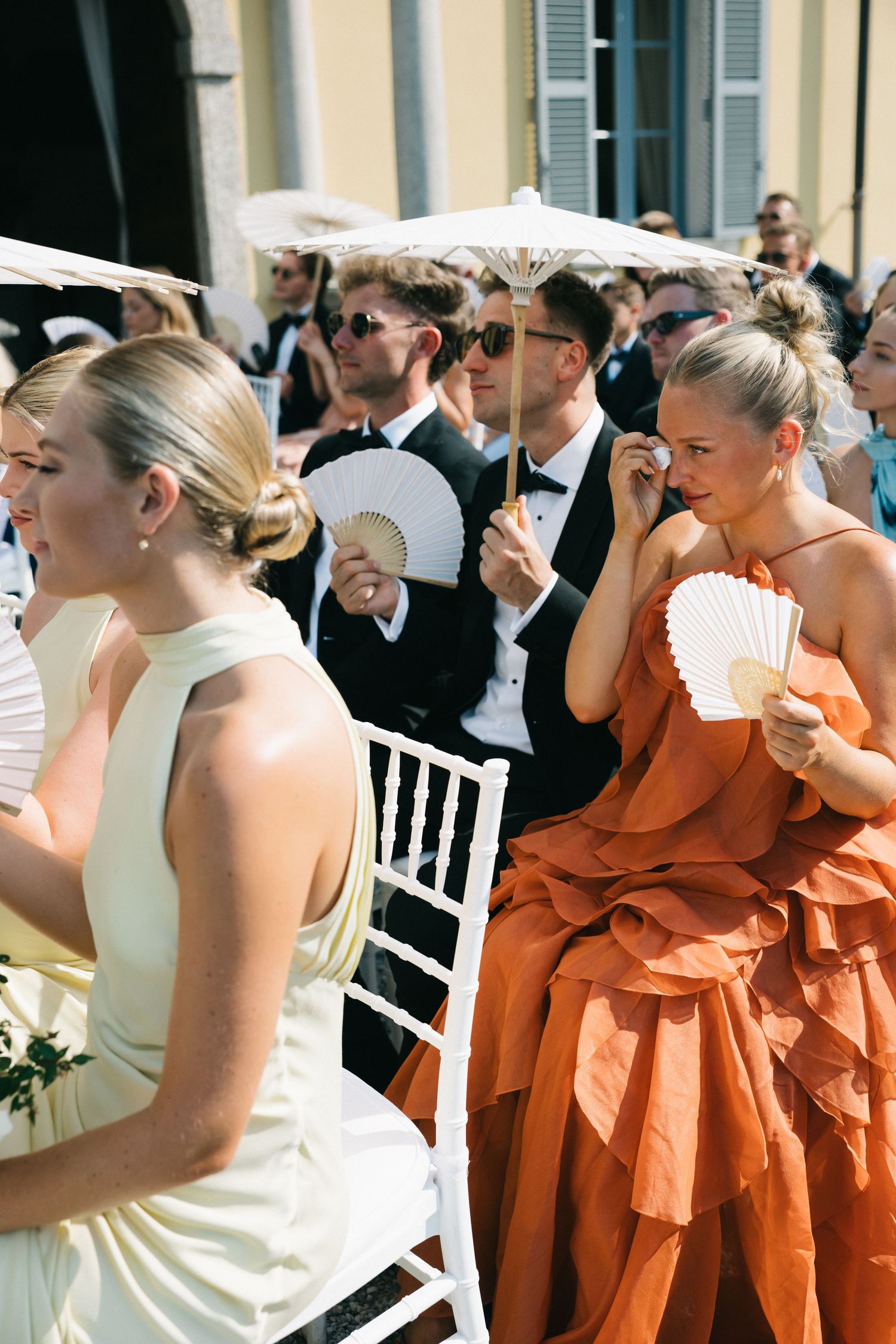 Elegant wedding guests in formal attire use hand fans outdoors, women in cream and orange gowns, men in tuxedos.