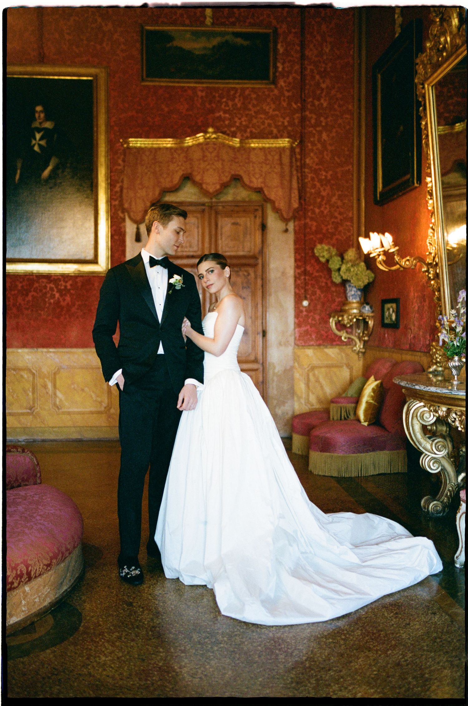 Elegant bride in white gown and groom in black tuxedo posing in ornate red-walled Italian wedding venue.