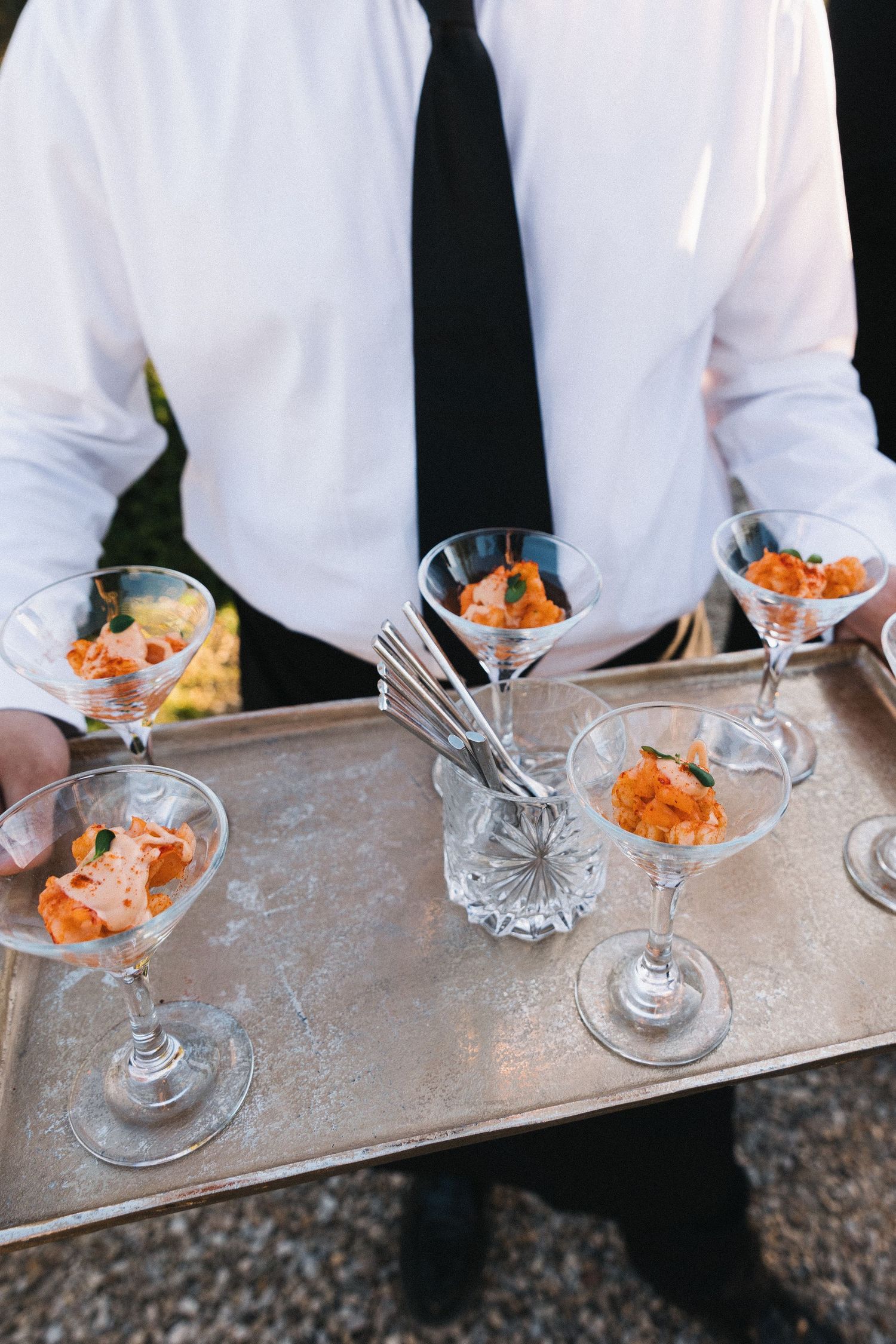 Waiter in white shirt and black tie serving orange appetizers in martini glasses on a silver tray.