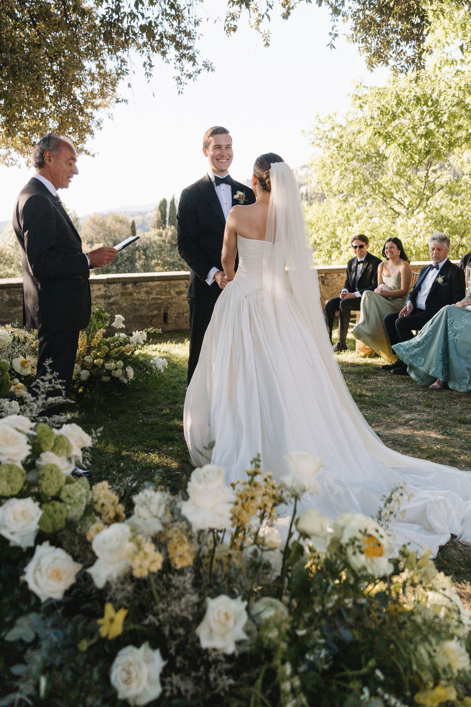 Bride and groom exchange vows at outdoor garden ceremony surrounded by white floral arrangements and seated guests.