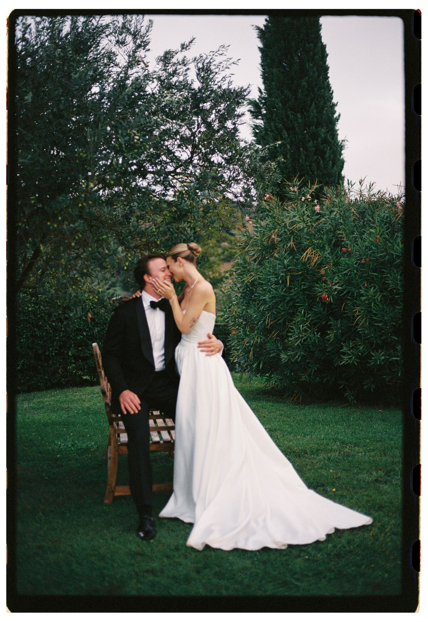 Bride and groom share romantic kiss in lush Italian garden with cypress trees on wedding day.