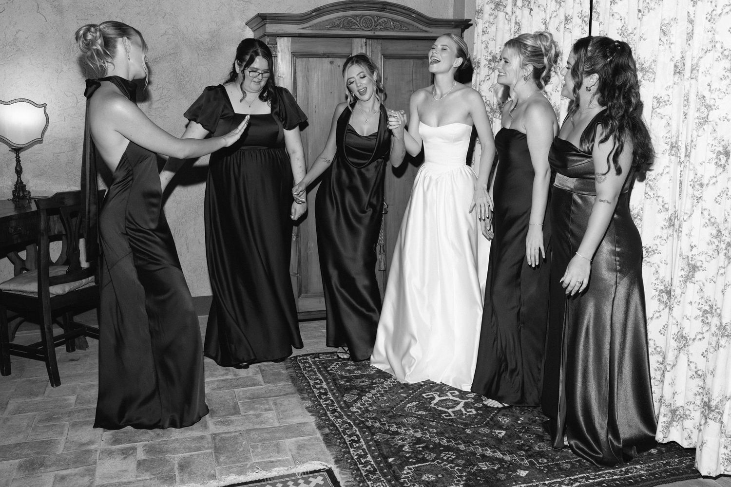 Black and white photo of a bride in white gown with bridesmaids in dark dresses celebrating together indoors.