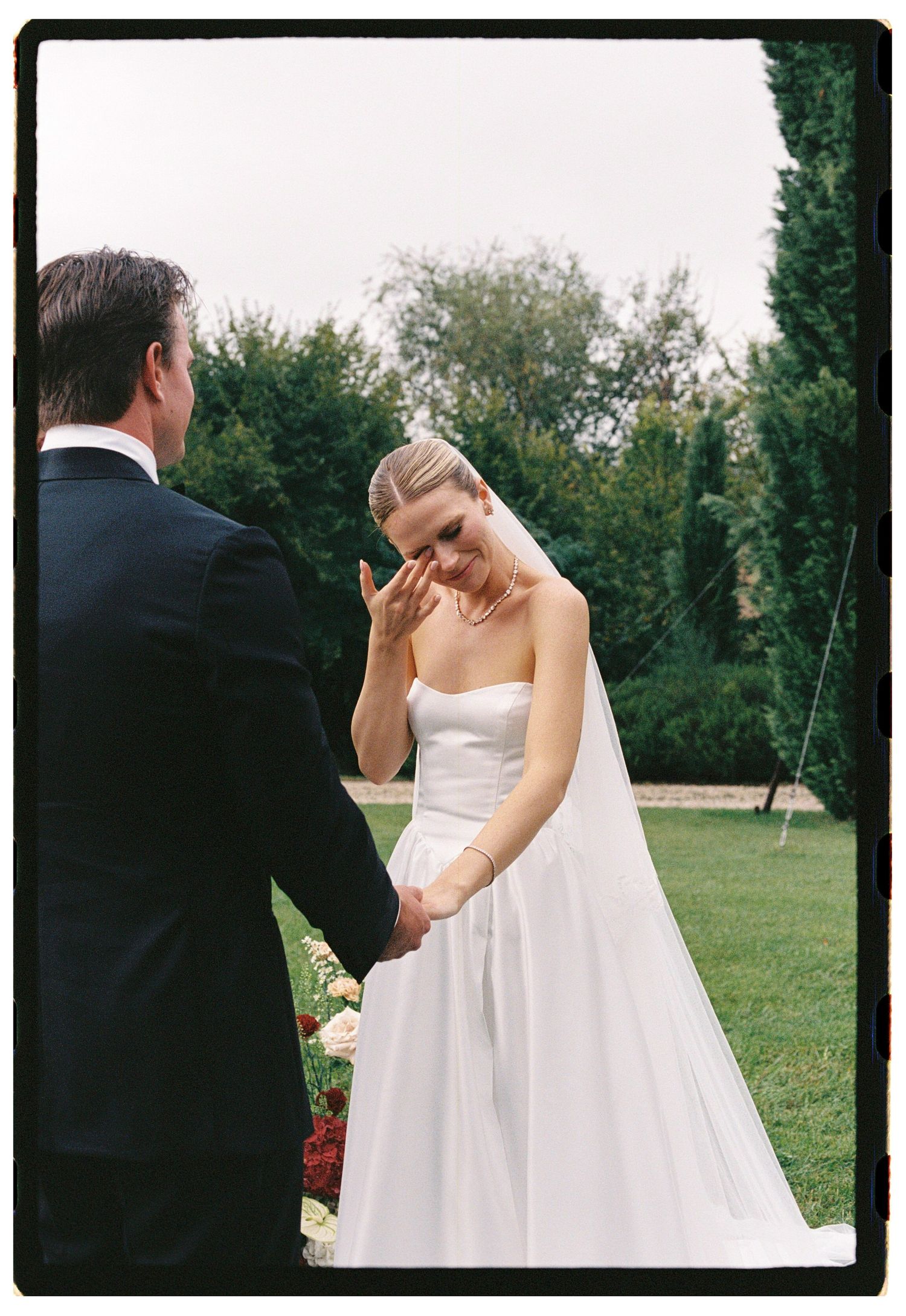 Emotional bride in strapless white gown wiping tears while holding hands with groom in black suit outdoors.