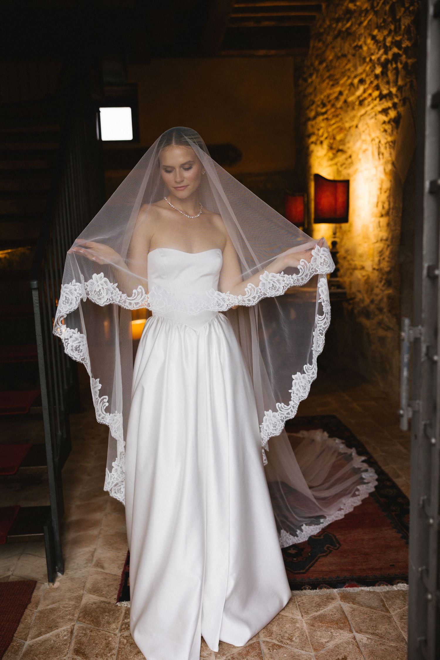 Bride in strapless white gown holding lace-trimmed veil in rustic stone venue with warm lighting.
