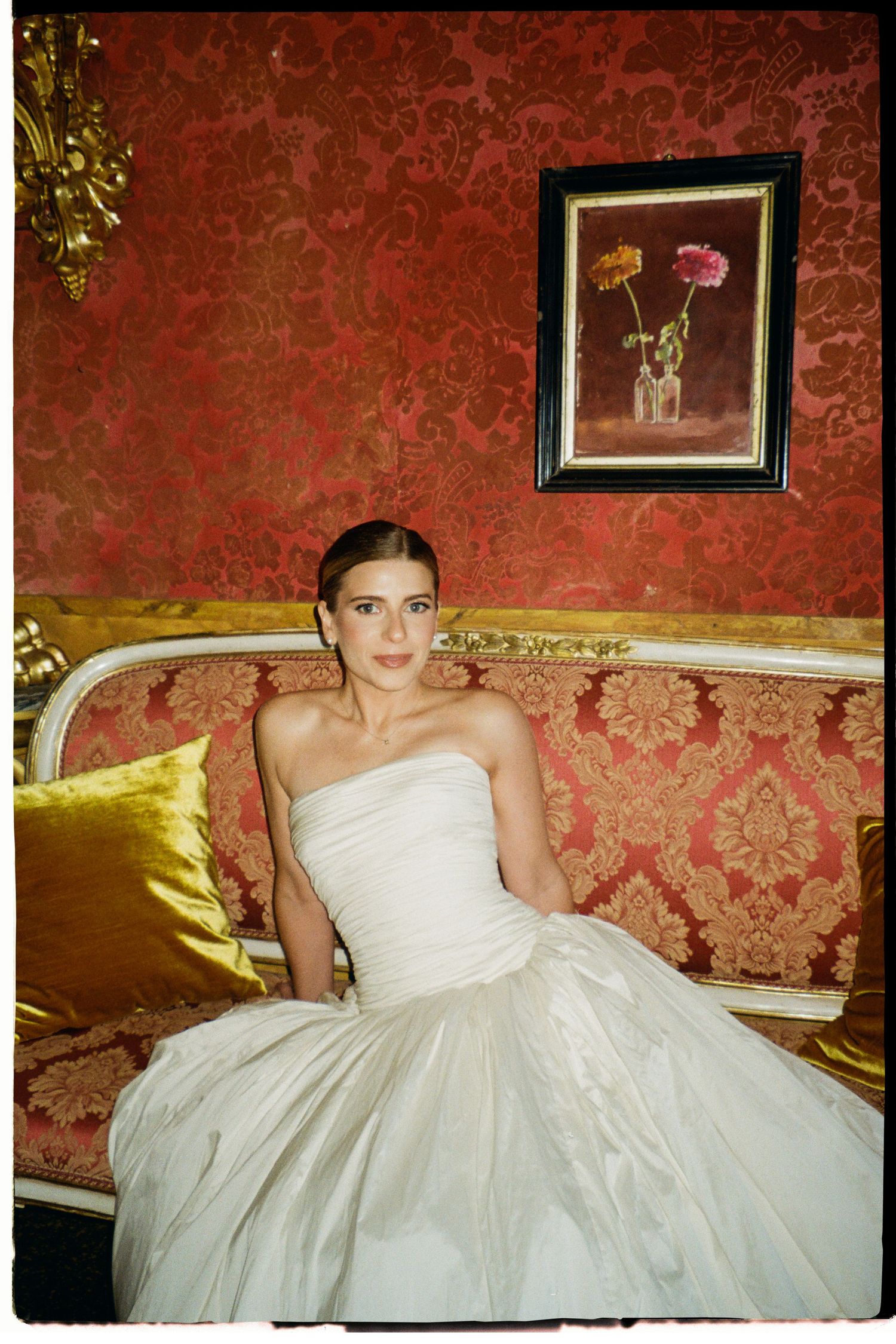 Elegant bride in strapless white ball gown seated on ornate gold sofa against red damask wallpaper.