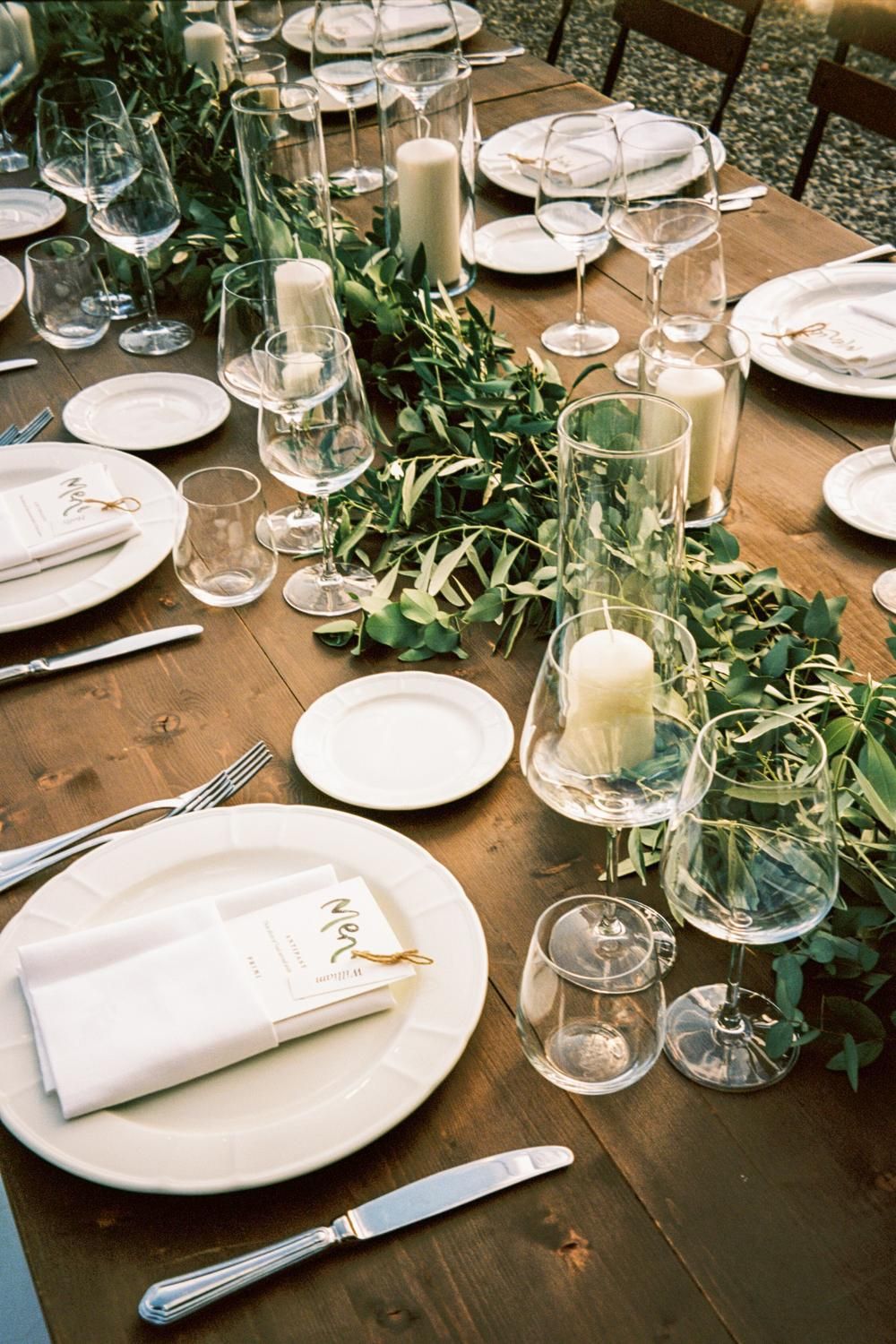 Elegant rustic wedding table setting with greenery garland, candles, and glassware.