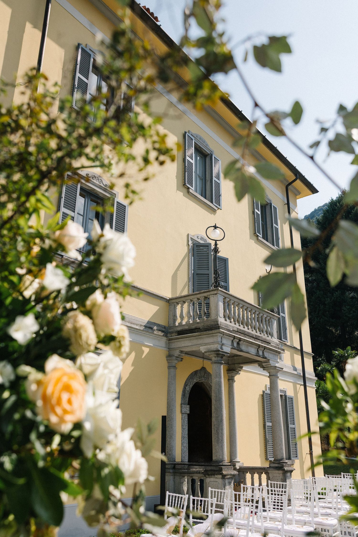Elegant yellow Italian villa with gray shutters, balcony, white roses in foreground, perfect wedding venue setting.