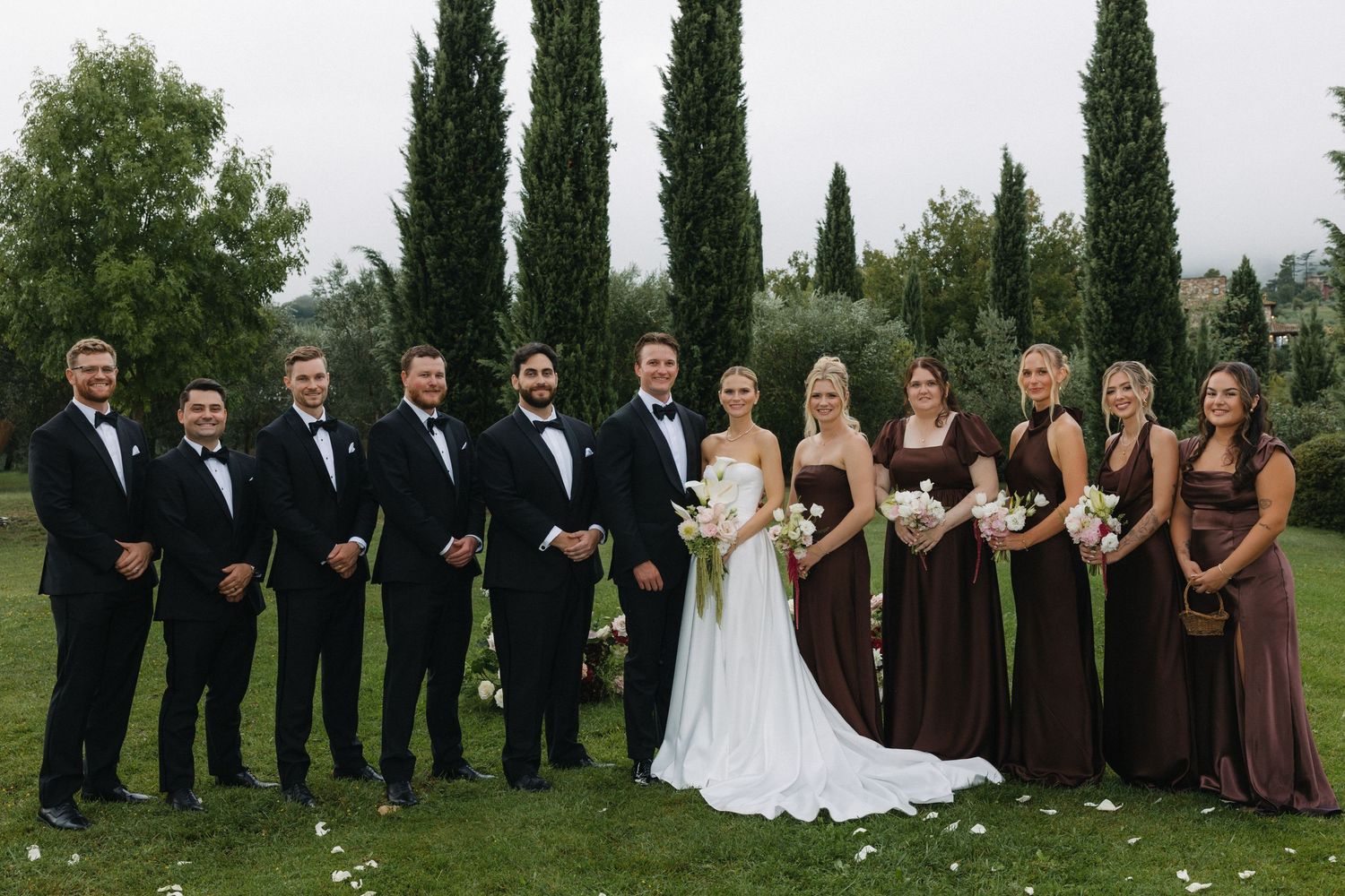 Wedding party poses outdoors with cypress trees, groomsmen in black tuxedos and bridesmaids in brown dresses flanking the bride and groom.