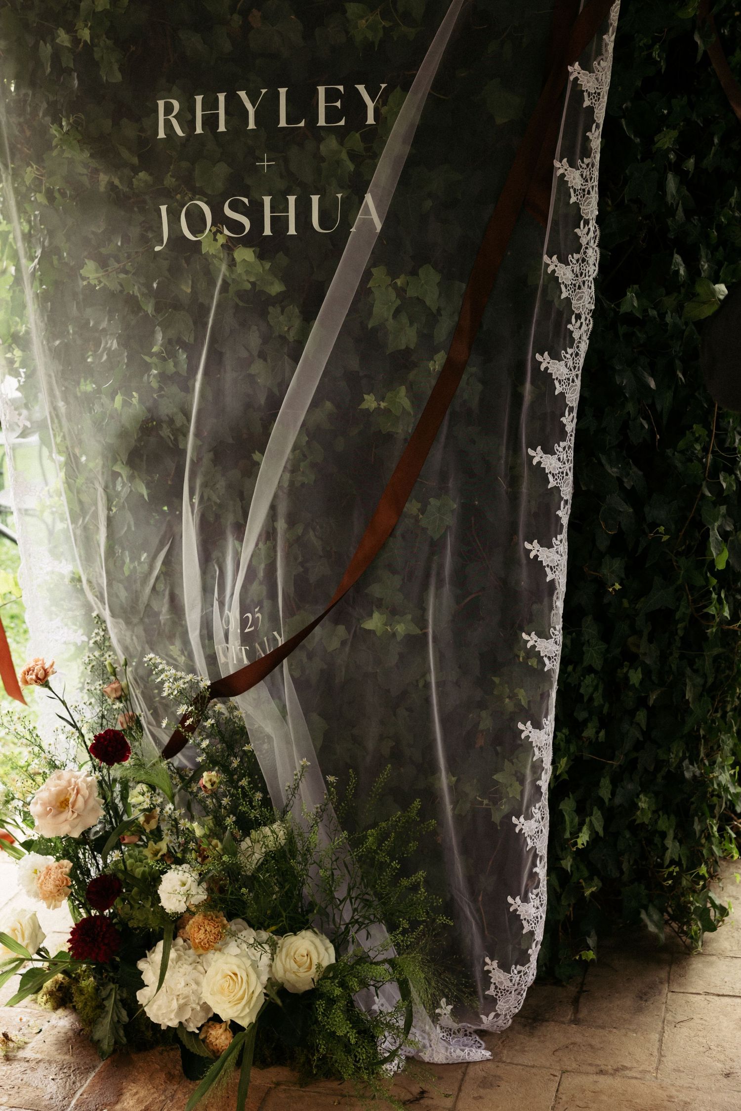 Elegant wedding backdrop with lace veil, greenery wall, floral arrangement, and Rhyley and Joshua signage.