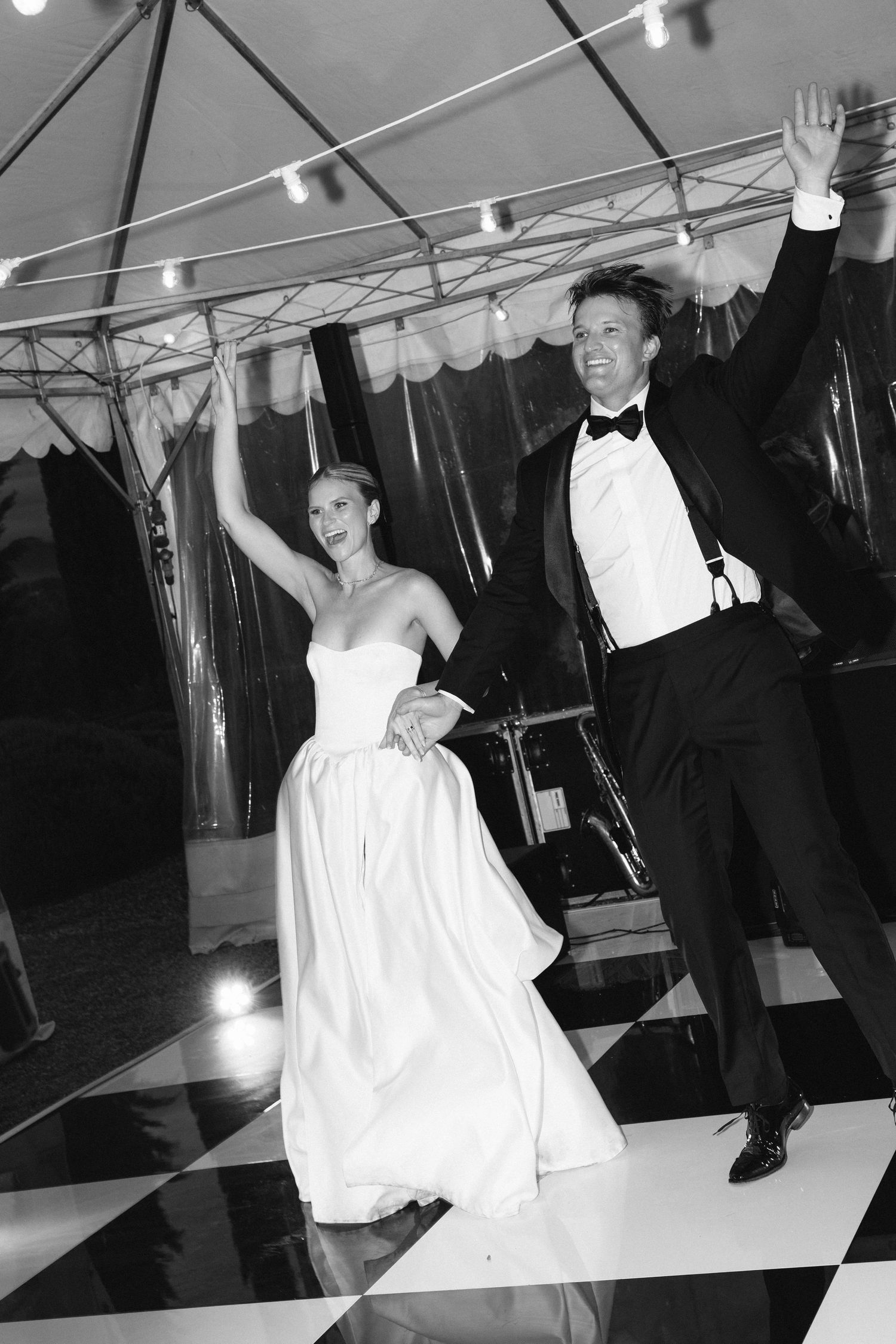 Black and white photo of bride and groom dancing joyfully under a tent on a checkered dance floor at their wedding reception.