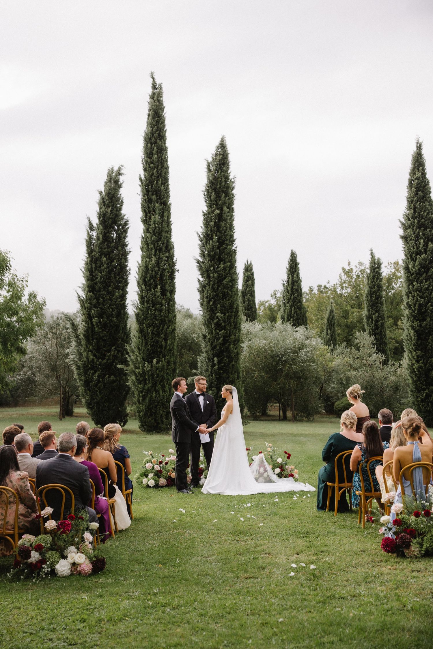 Outdoor wedding ceremony on green lawn with tall cypress trees, bride and groom exchange vows before seated guests.