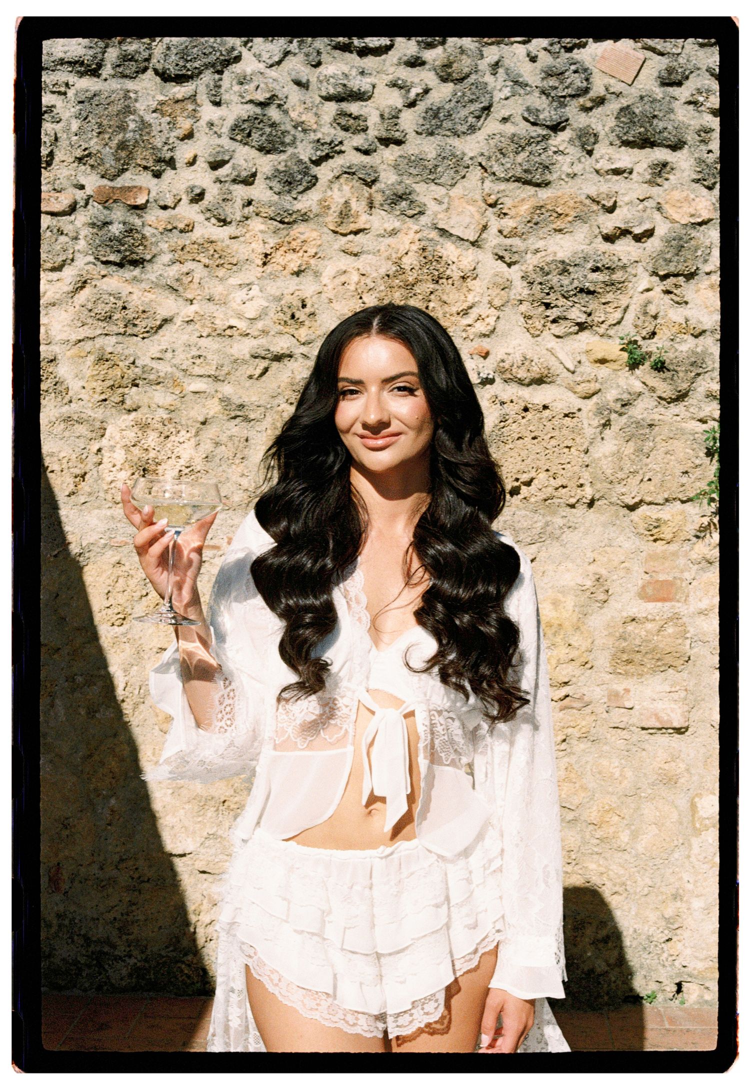 Woman in white lace crop top and shorts holding a champagne glass, posing against a rustic stone wall.