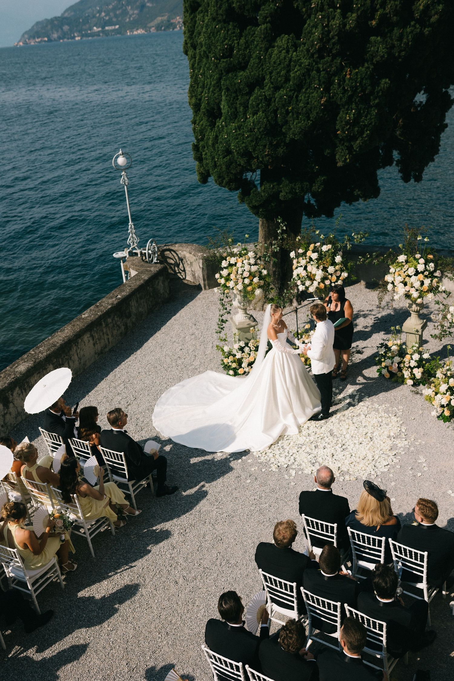 Elegant outdoor wedding ceremony on a lakeside terrace with floral arch, bride in white gown, guests seated in white chairs.