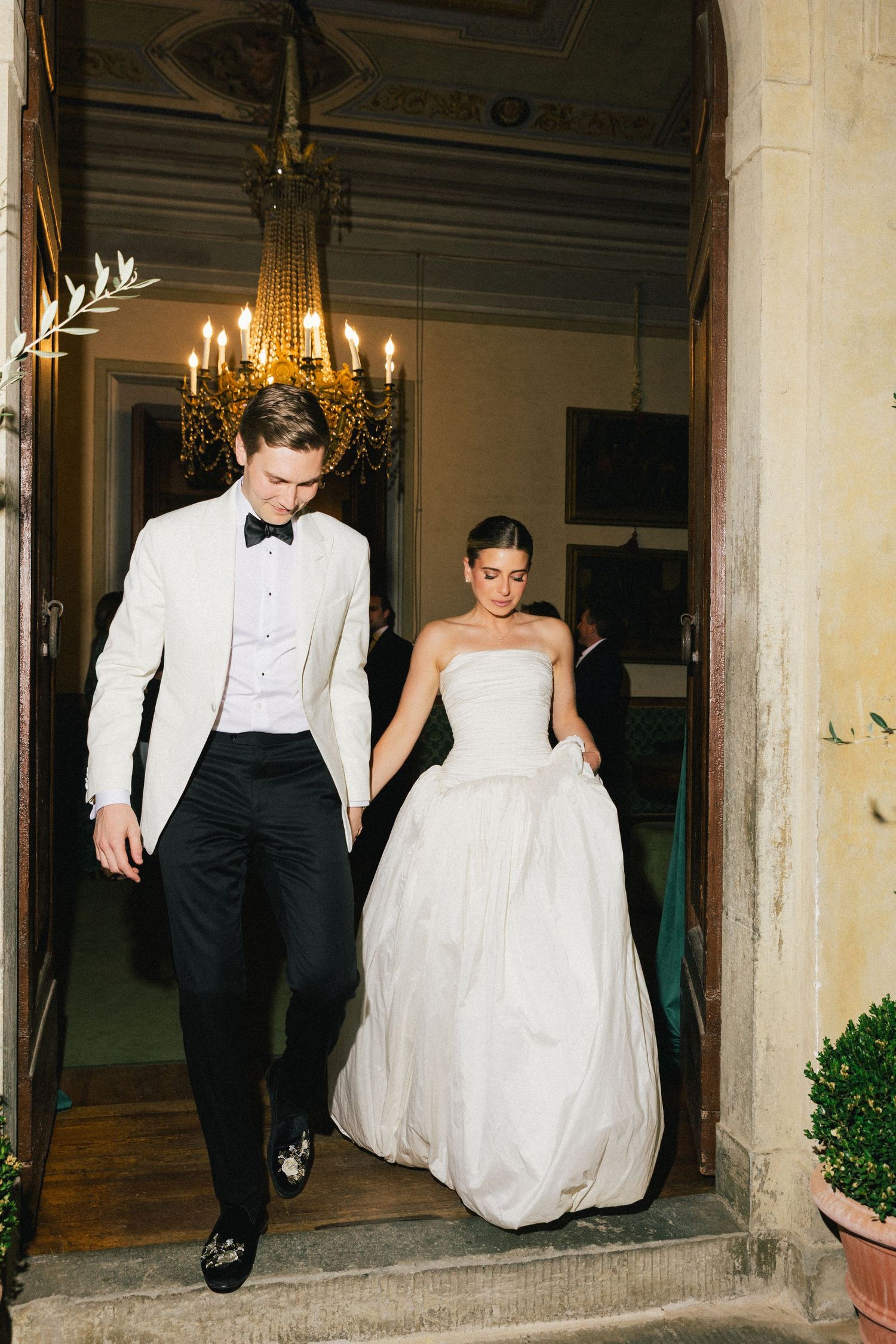 Bride and groom exit elegant venue, groom in white tuxedo jacket, bride in strapless ball gown, chandelier glowing behind them.
