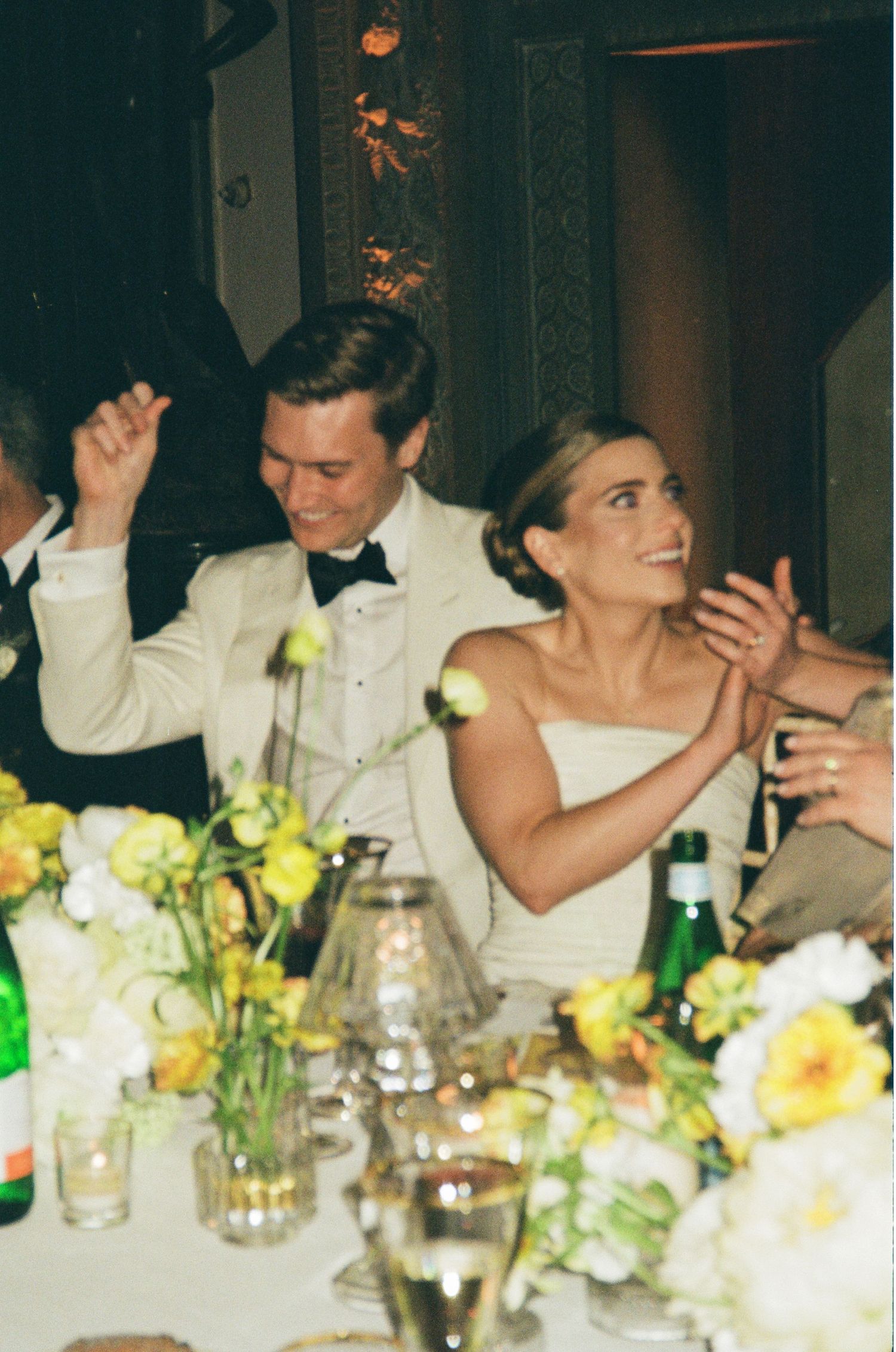 A joyful bride and groom in formal attire celebrate at their wedding reception surrounded by yellow floral centerpieces.