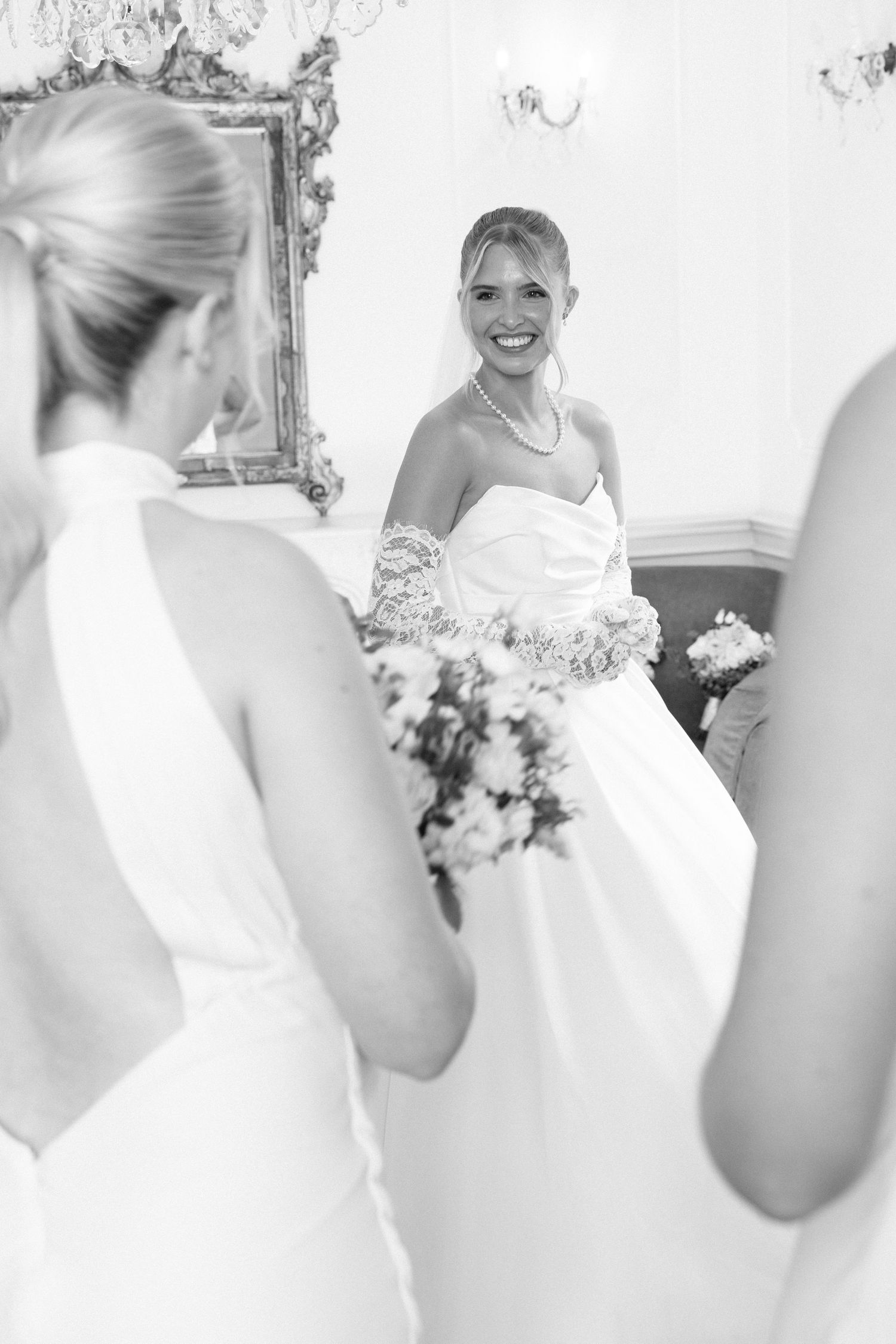 Smiling bride in strapless wedding gown holding bouquet, reflected in ornate mirror, black and white photo.