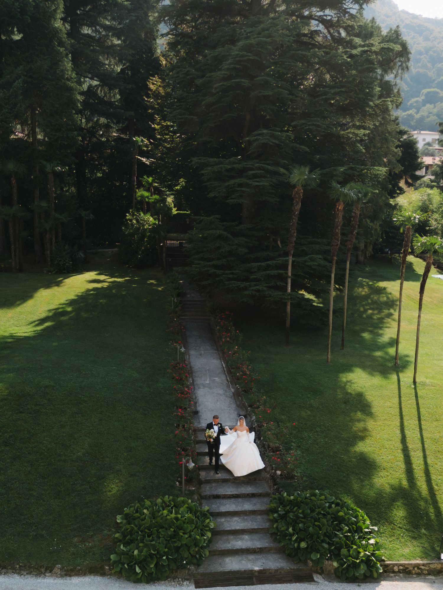Aerial view of a bride and groom posing on garden steps surrounded by lush green trees at an elegant wedding venue.