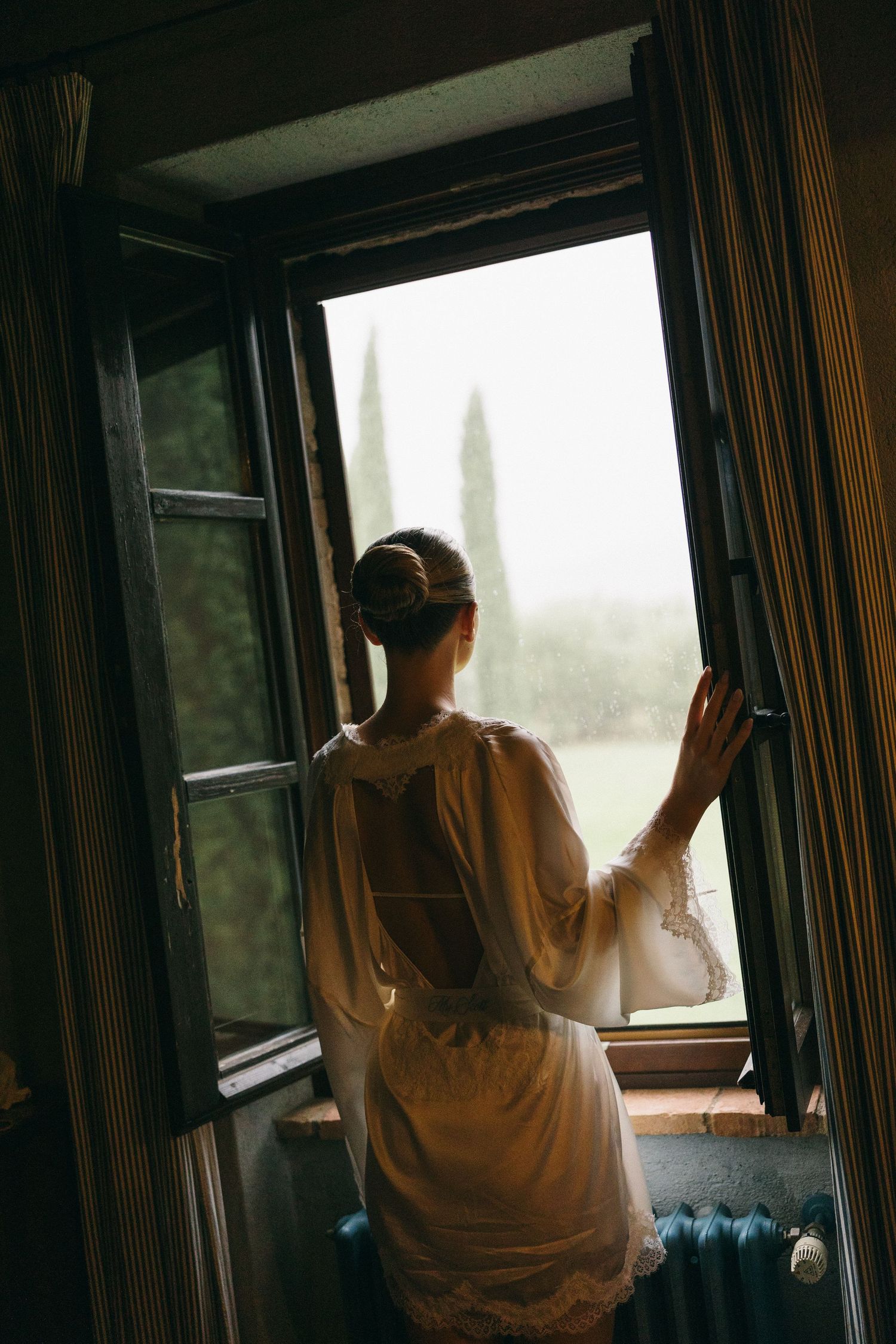 Woman in white lace robe gazing out open vintage window at cypress trees in Tuscany landscape.