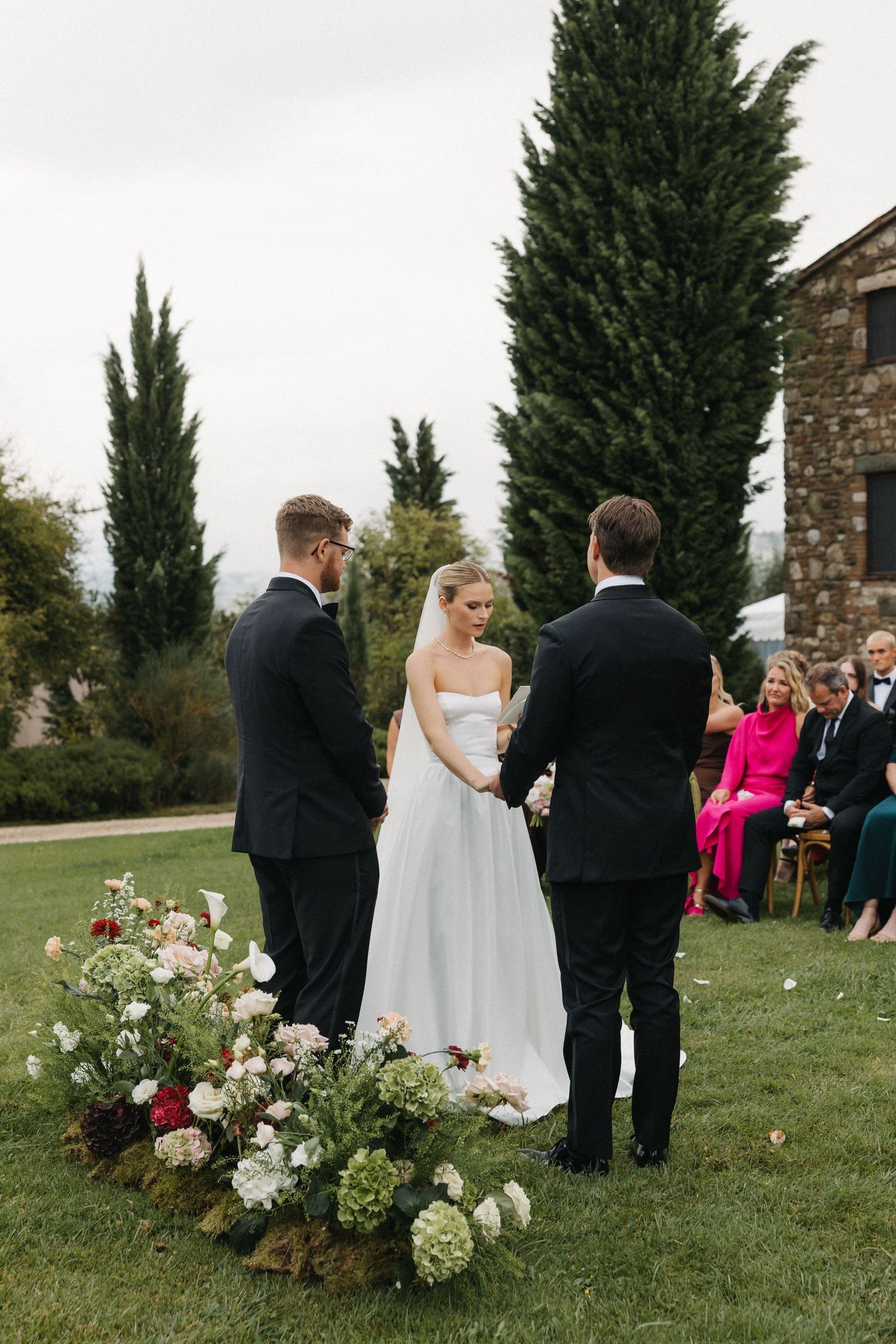 Outdoor wedding ceremony on green lawn with bride in white gown and groom in black tuxedo, flanked by floral arrangements.