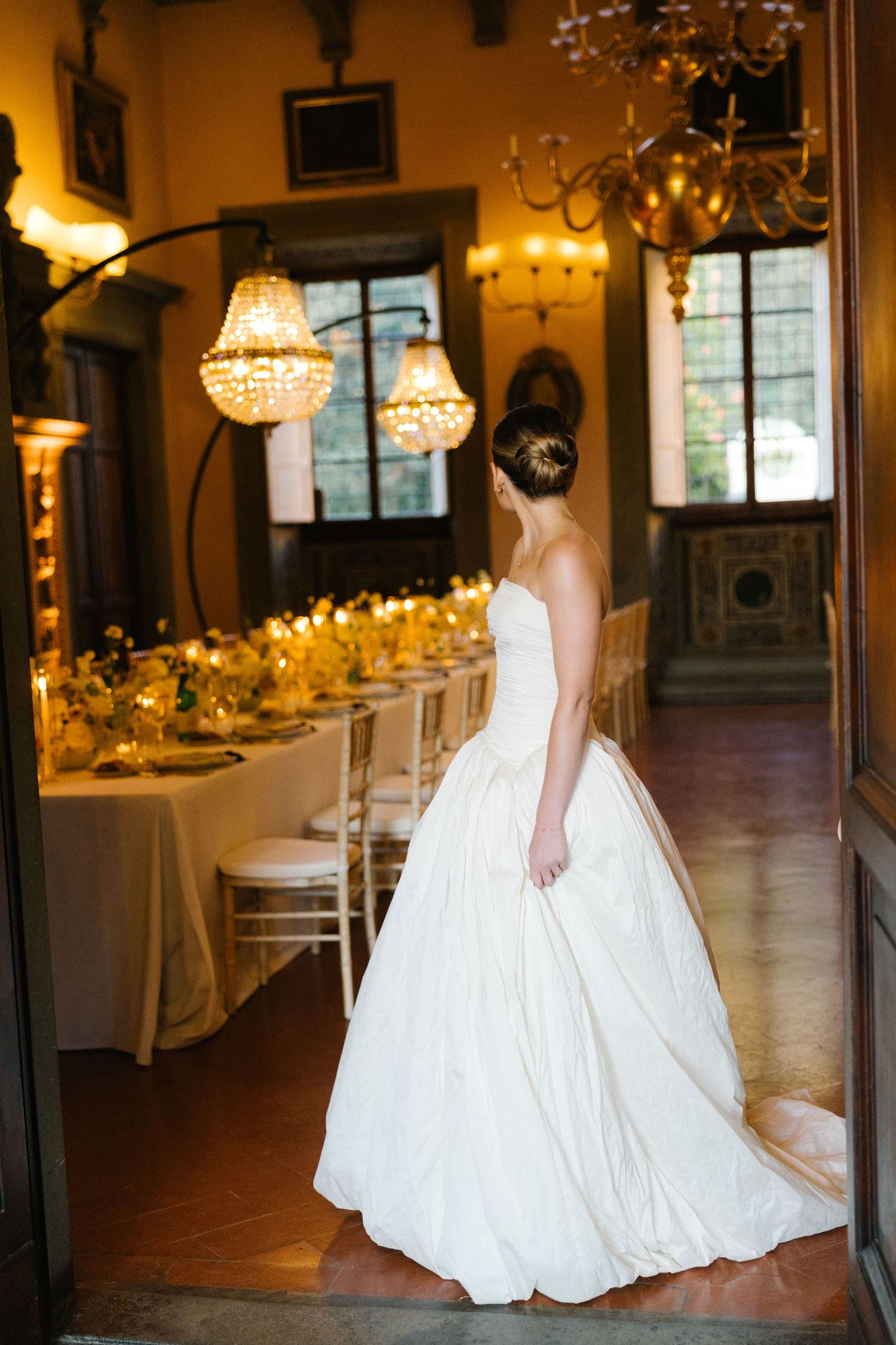 Bride in strapless white ballgown stands in doorway of elegantly lit wedding reception hall with crystal chandeliers.