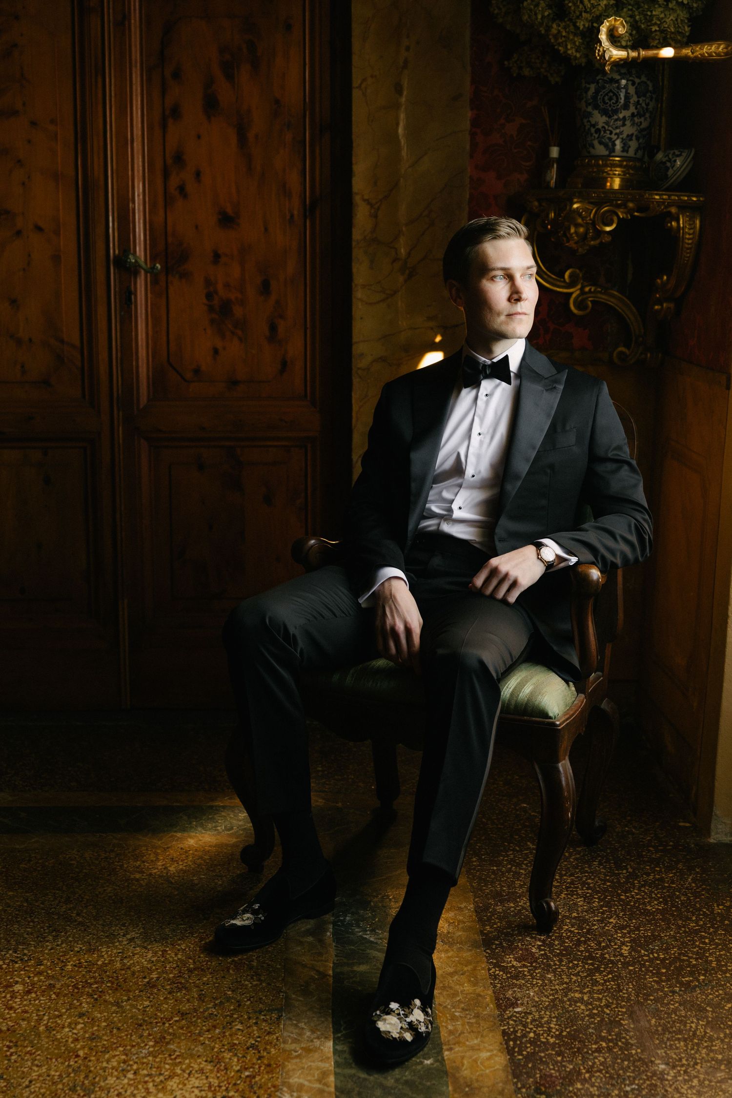 Elegant groom in black tuxedo with bow tie seated in ornate chair in a luxurious dimly lit room.