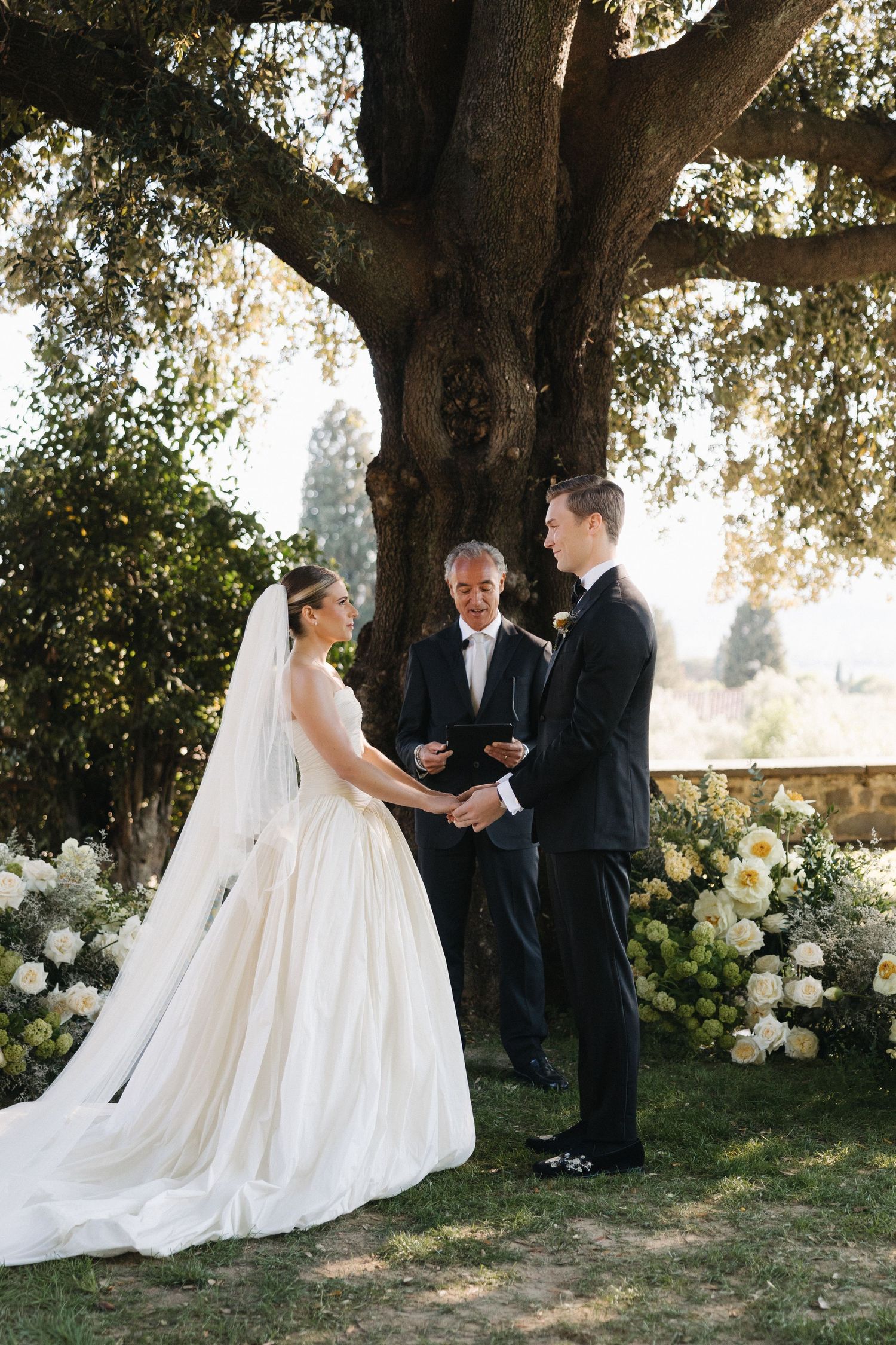 Bride and groom exchange vows outdoors under a large tree with white floral arrangements at garden wedding.