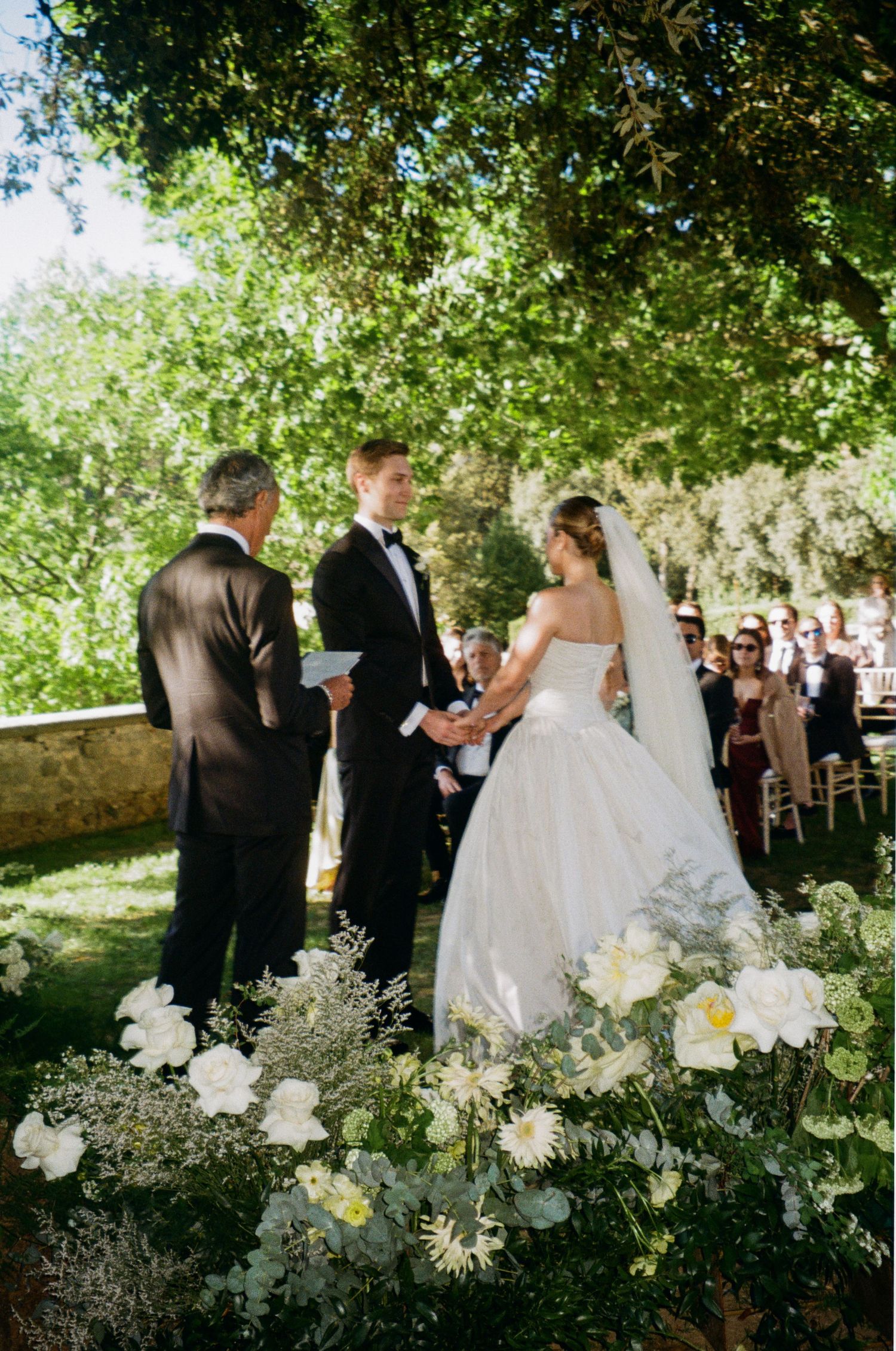 A bride and groom hold hands during an outdoor garden wedding ceremony surrounded by white wildflowers and guests.