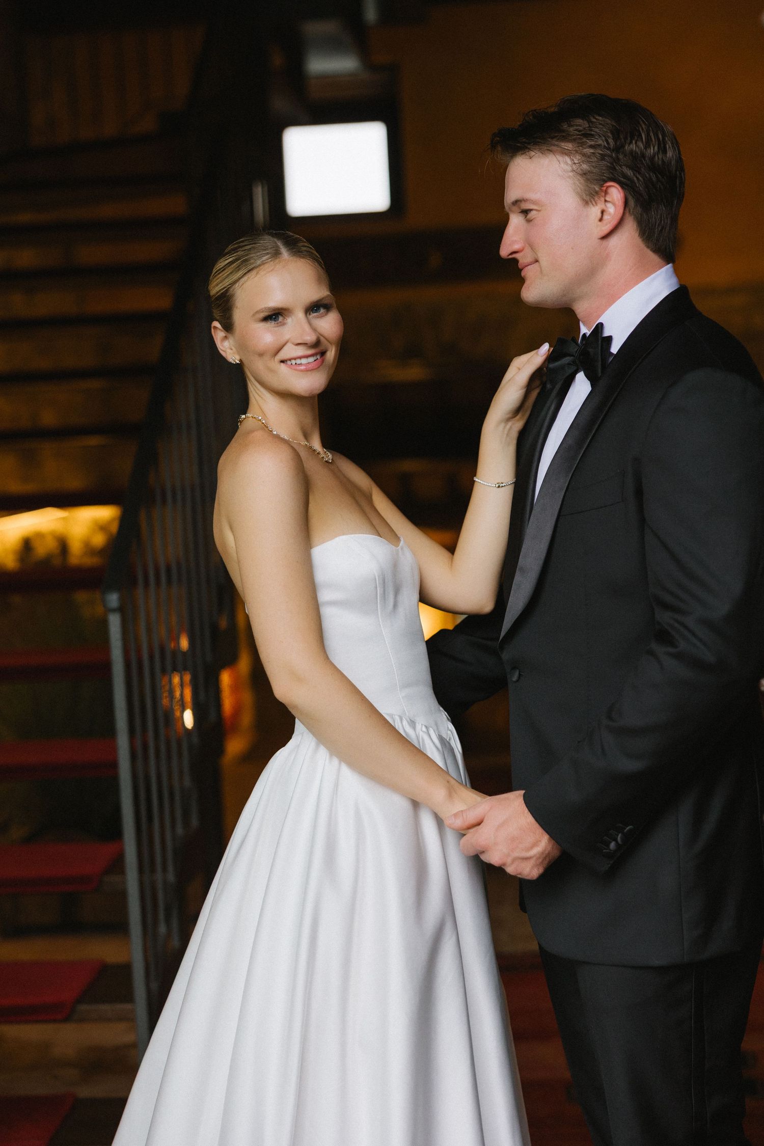 Bride in white strapless gown and groom in black tuxedo share a romantic moment on staircase at wedding reception.