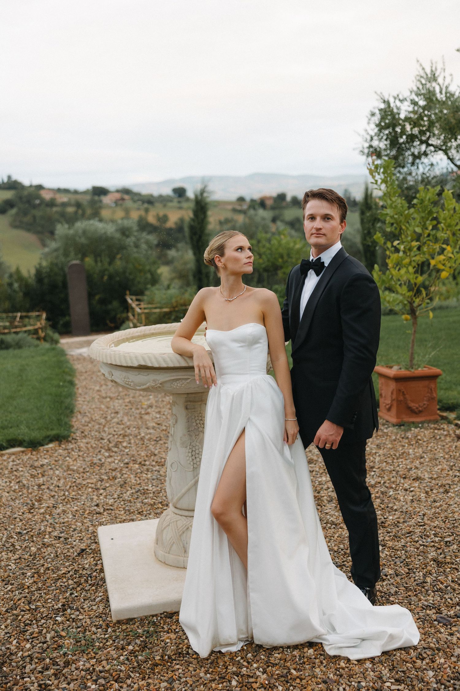 Bride in white strapless gown with slit and groom in black tuxedo posing by stone fountain in Tuscan garden.