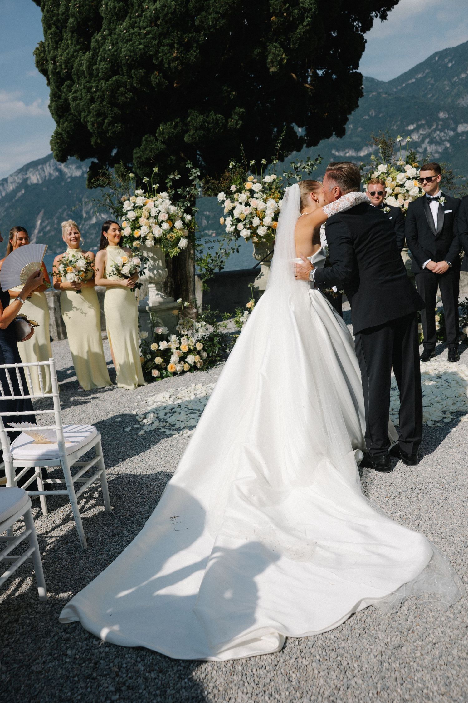 Bride and groom share first kiss at outdoor lakeside wedding ceremony with bridesmaids and mountain views.