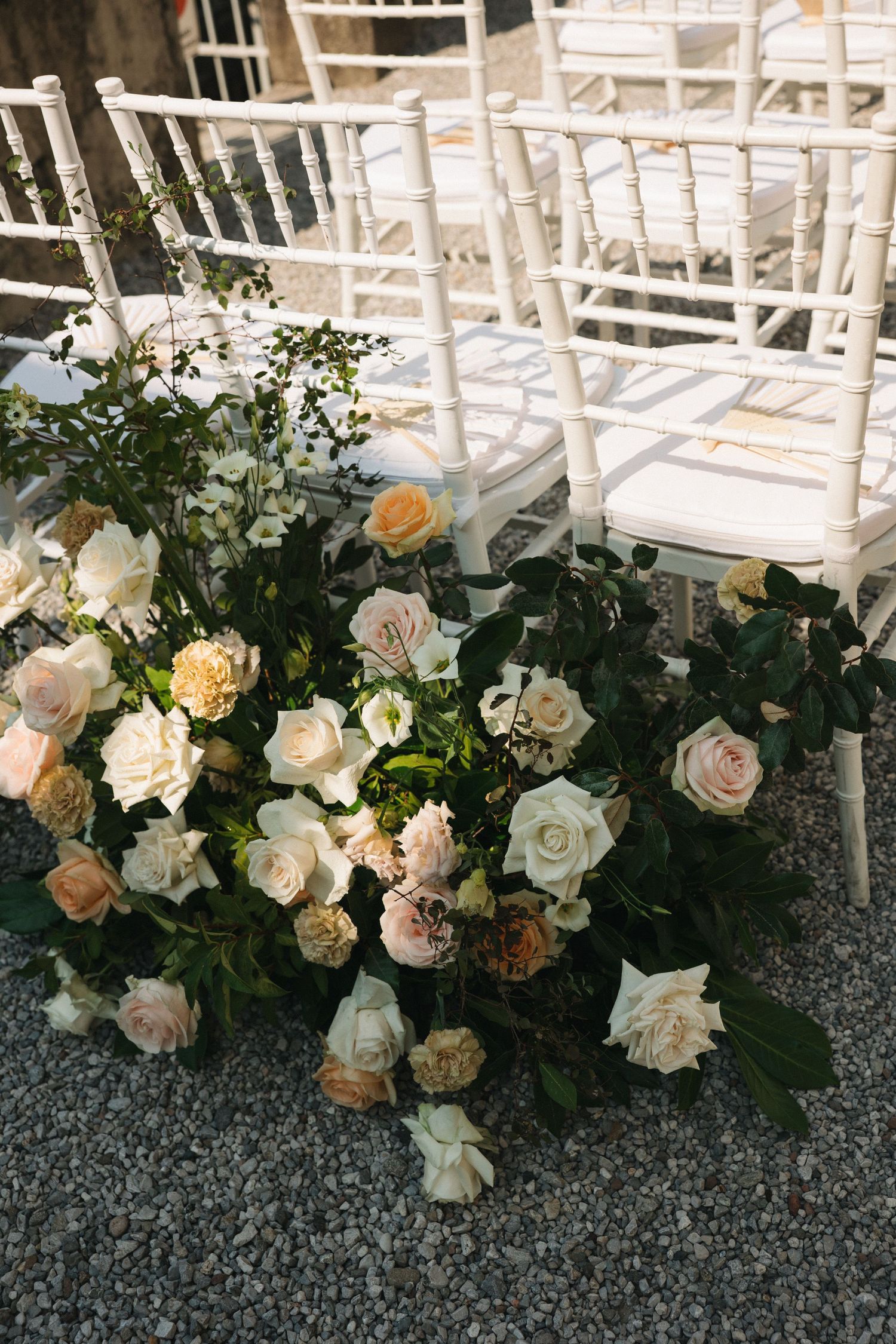 Elegant wedding ceremony setup with white Chiavari chairs and lush floral arrangements featuring blush and cream roses on gravel.