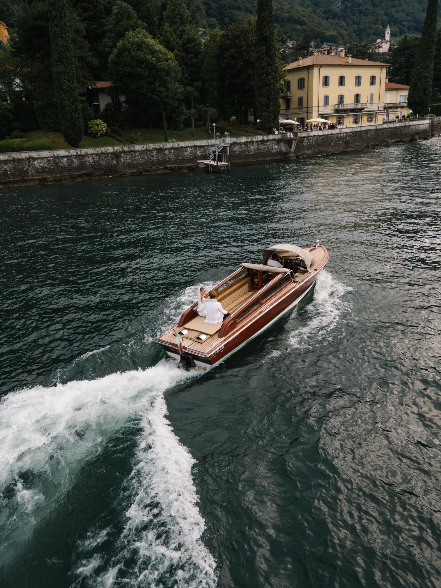 A classic wooden speedboat cruises along a scenic European lake with a yellow villa and lush trees in the background.
