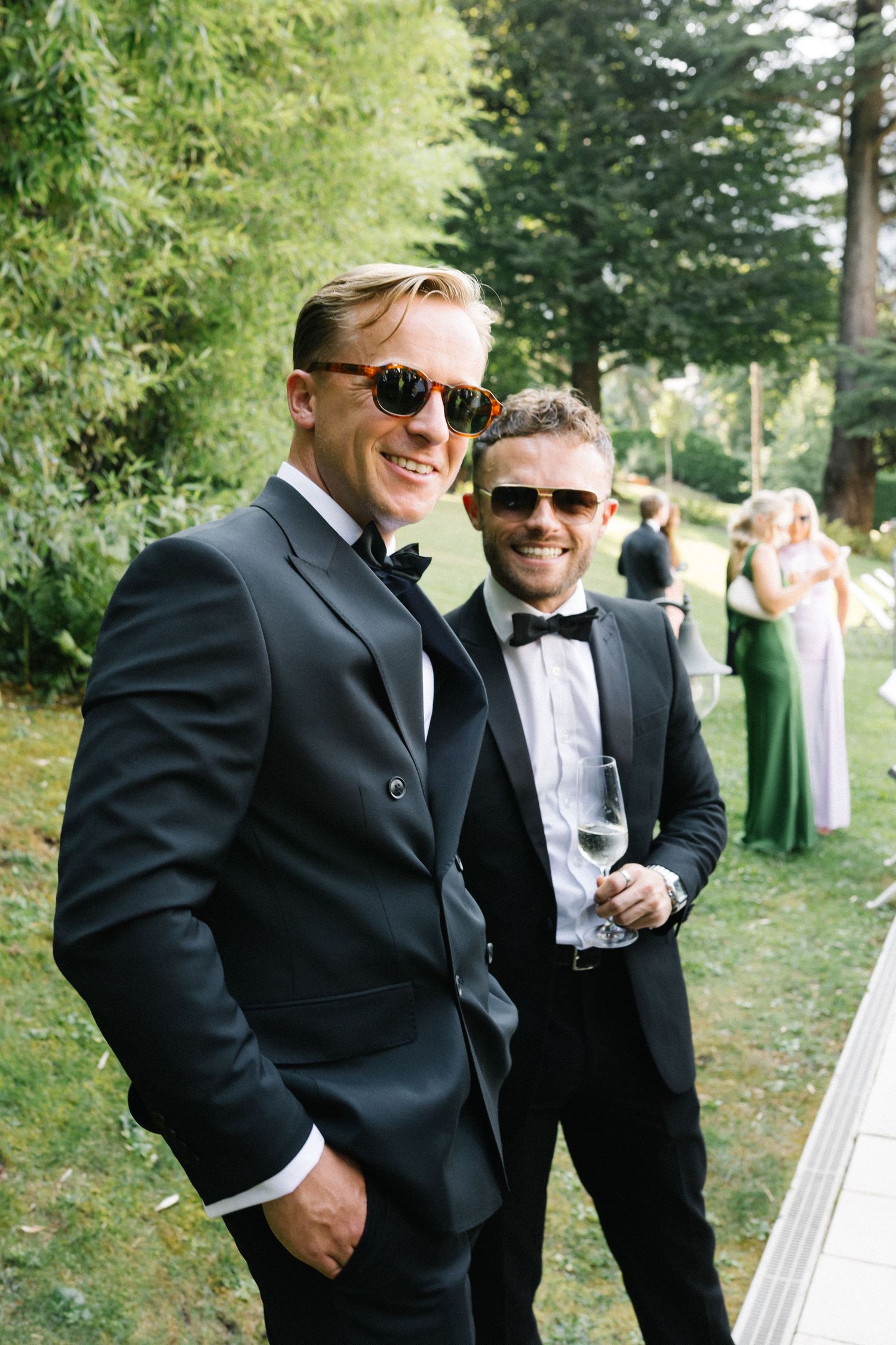 Two men in black tuxedos and sunglasses smile at a garden wedding reception with guests in background.