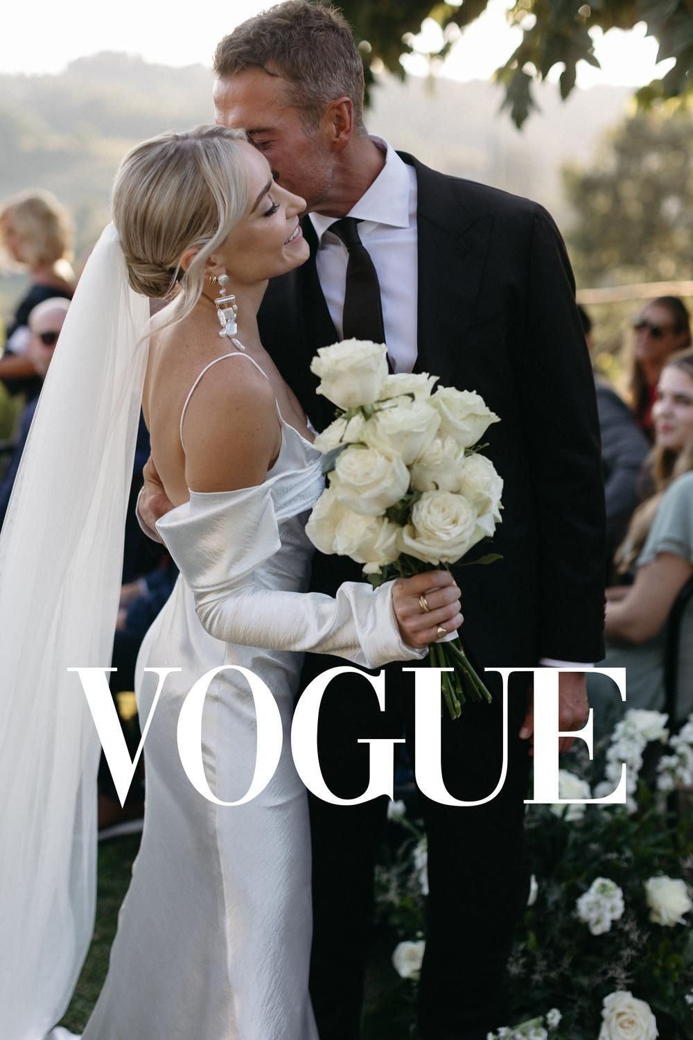 Vogue featured wedding with blonde bride holding white roses bouquet sharing kiss with groom at outdoor ceremony.