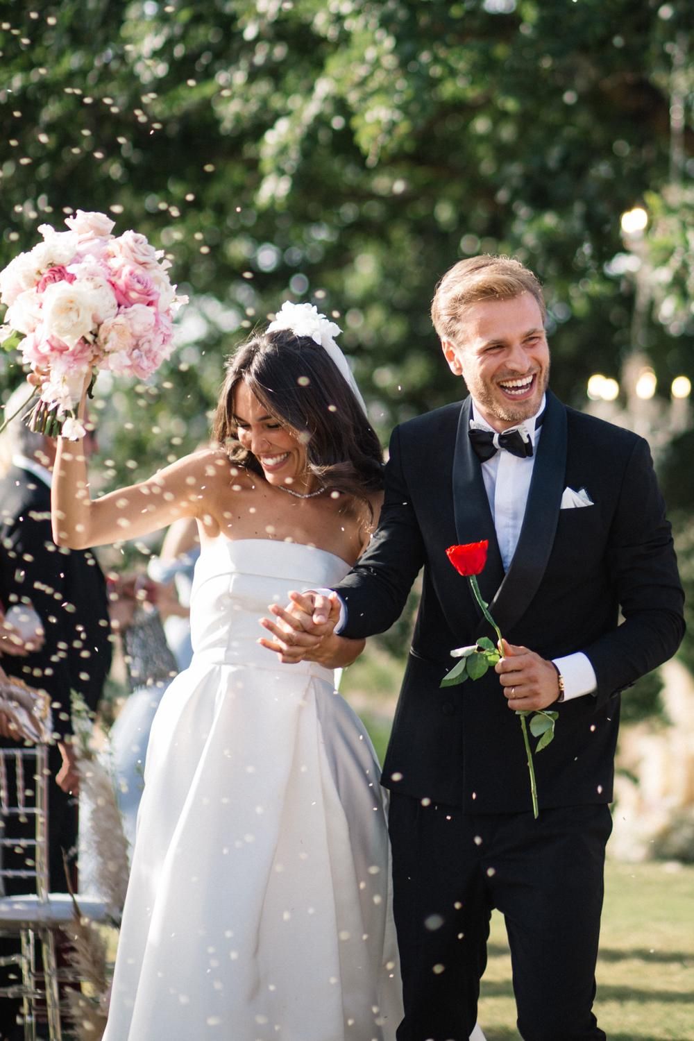 Joyful newlyweds walking down aisle with confetti, bride holding pink bouquet, groom with red rose.