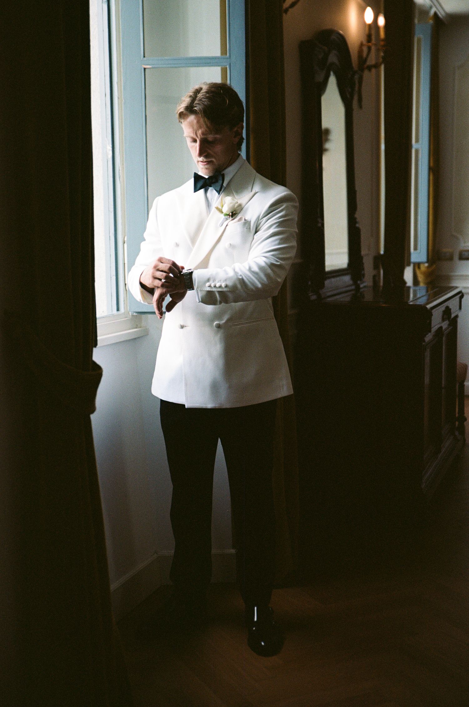 Groom in white double-breasted tuxedo jacket with bow tie and boutonniere adjusting cufflinks by a window.